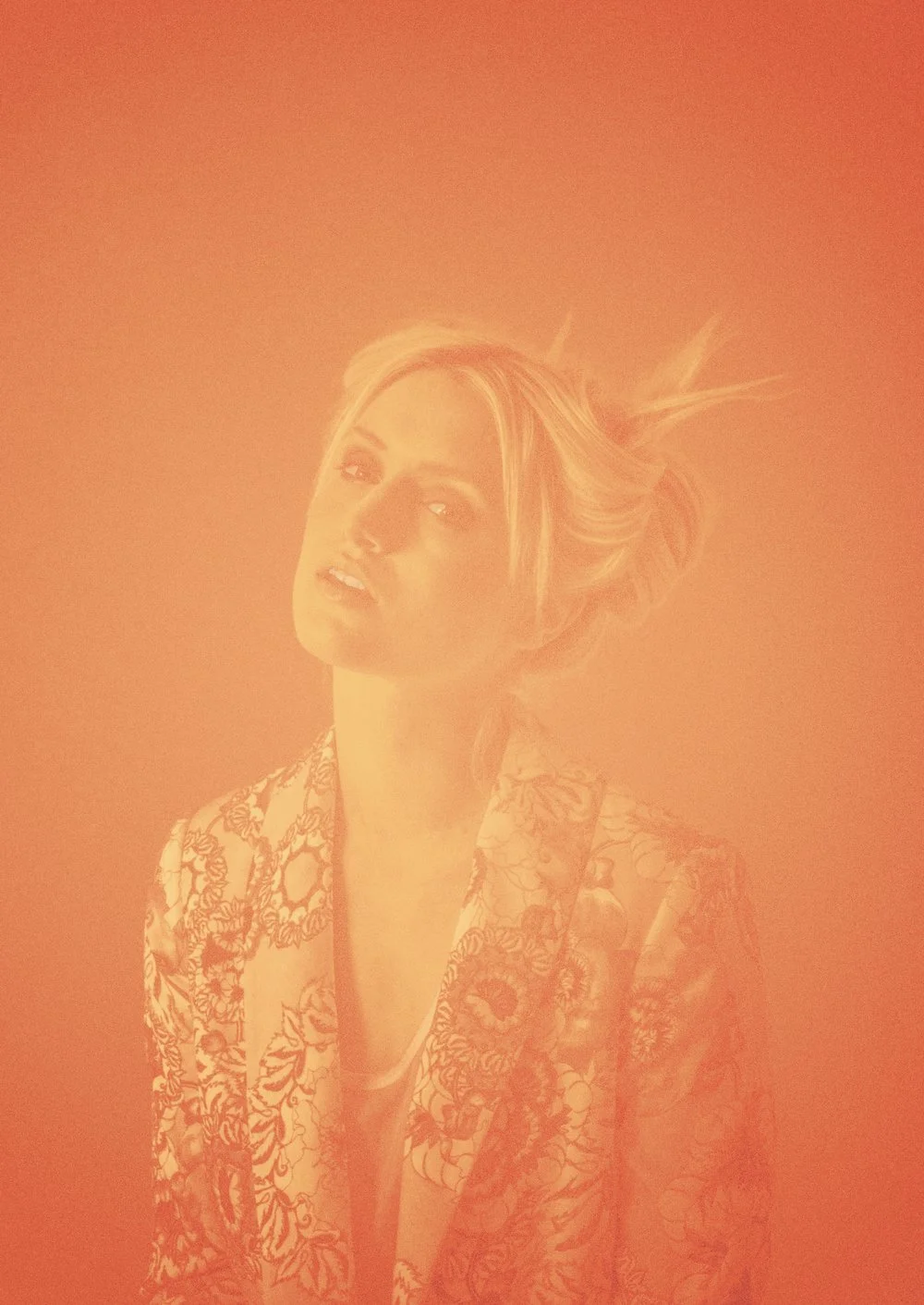 A woman with blonde hair in a loose updo, wearing a patterned jacket, gazes towards the camera against an orange background.