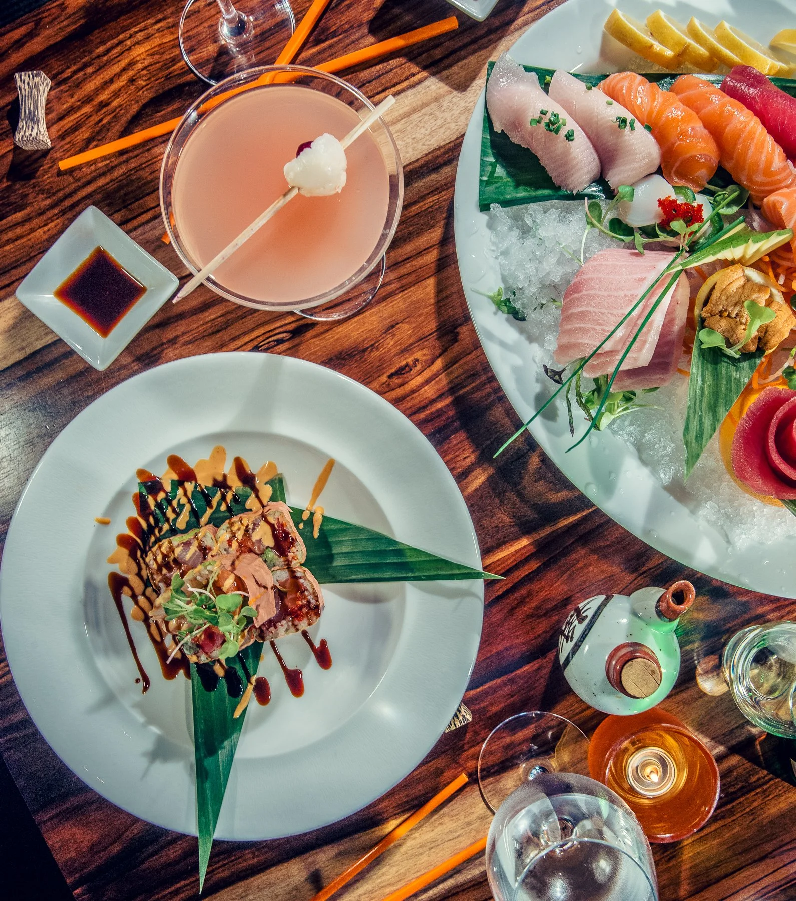 A table with a variety of food including sashimi, sushi rolls, a cocktail with a skewer, soy sauce, and glasses of wine or champagne.