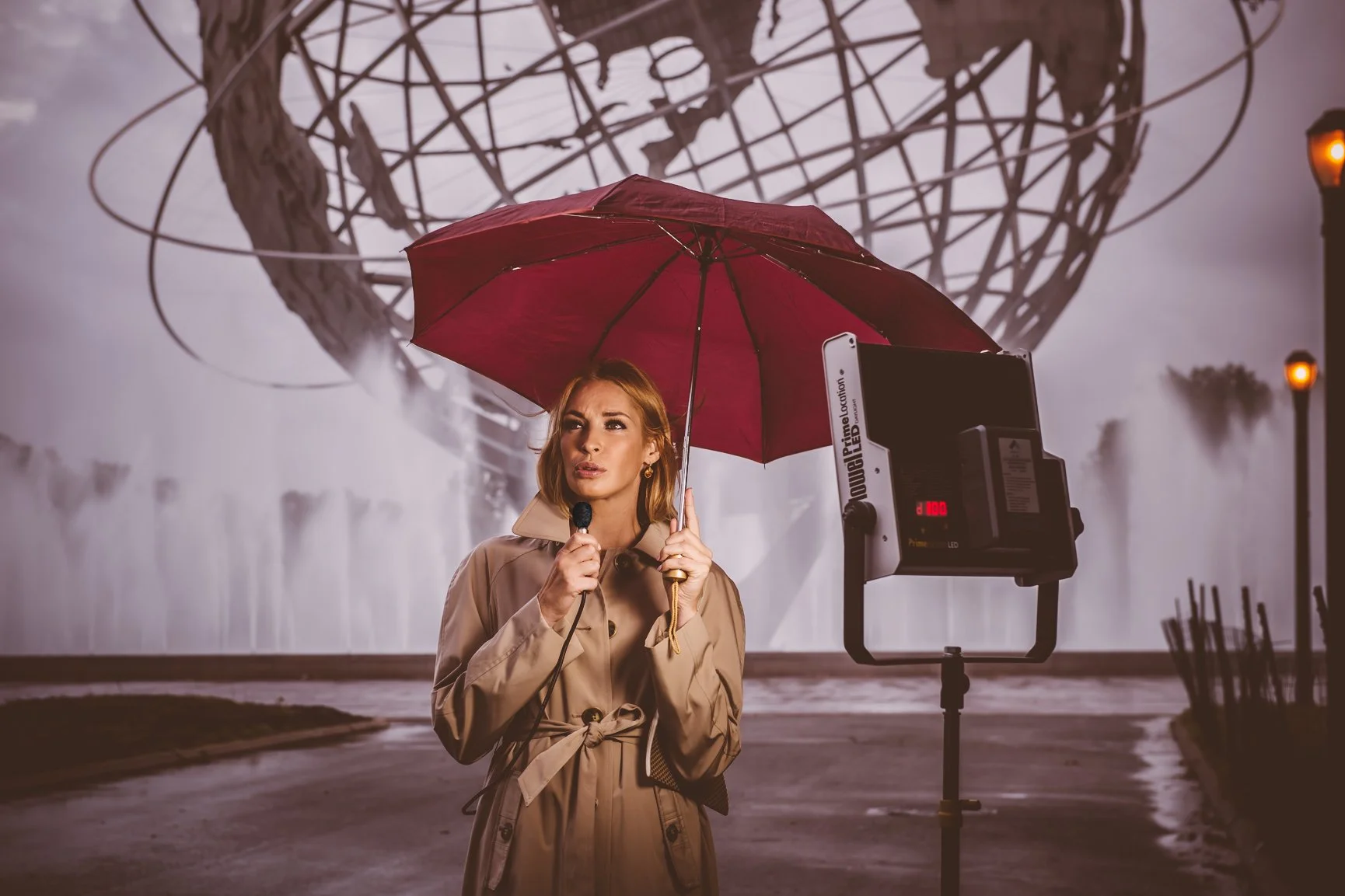 A woman holding a red umbrella and speaking into a microphone in front of a news camera at a location with a large globe sculpture and waterfalls in the background.