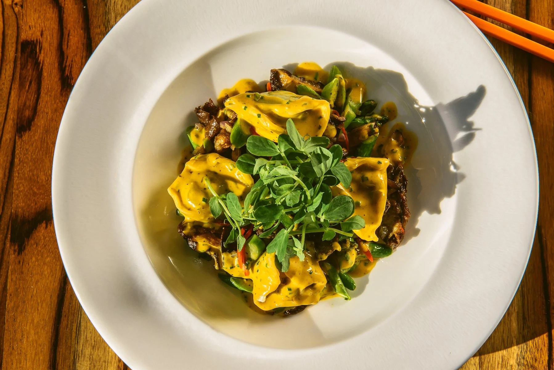 A white plate with a serving of Asian-style dumplings topped with fresh green herbs, served on a wooden table with orange chopsticks on the side.