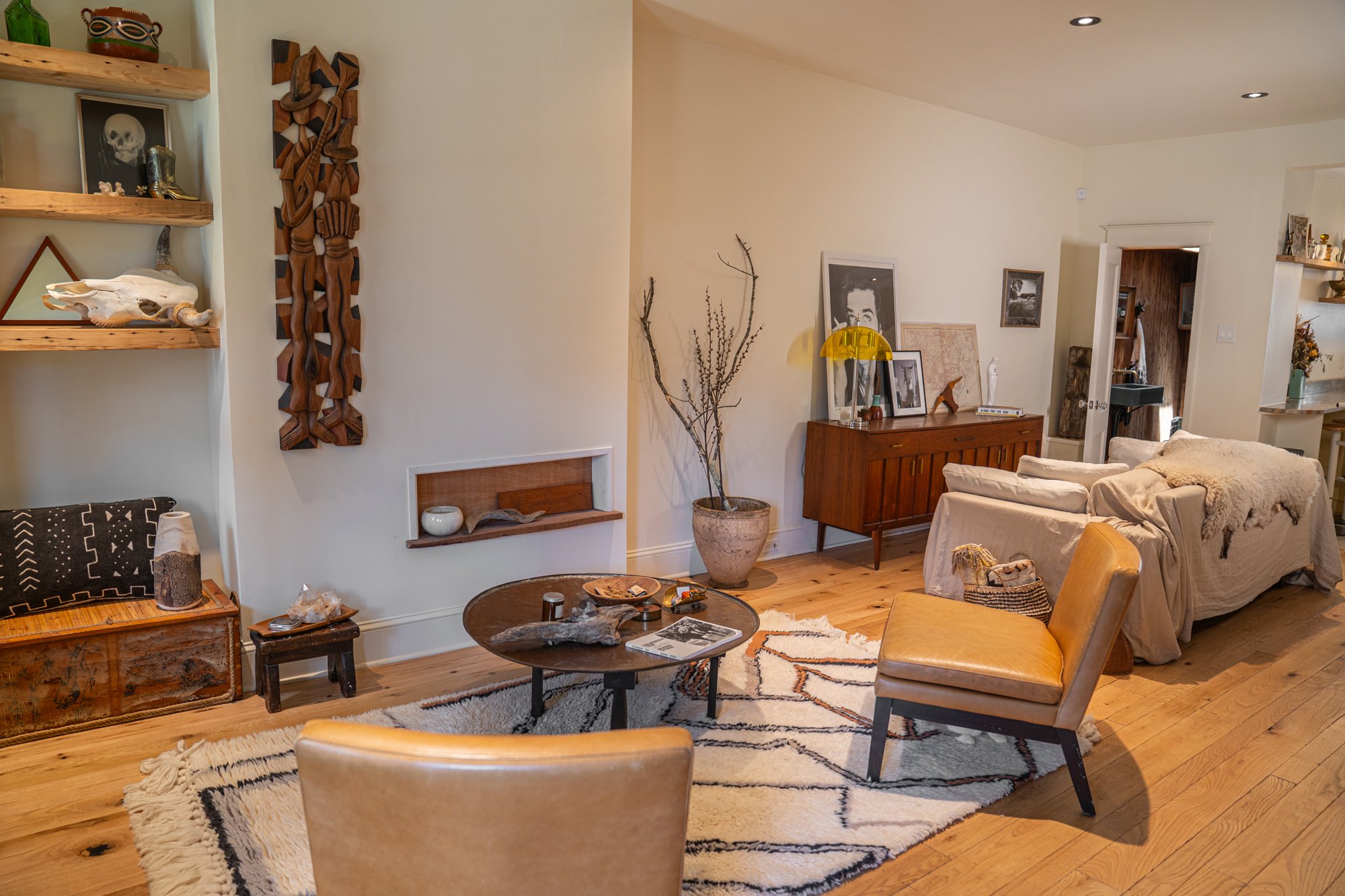 A cozy living room with wooden flooring, a patterned area rug, tan chairs, a white sofa covered with a throw, a round coffee table with decorative items, a wooden sideboard with framed photos and a yellow lamp, and various art and decorative objects 