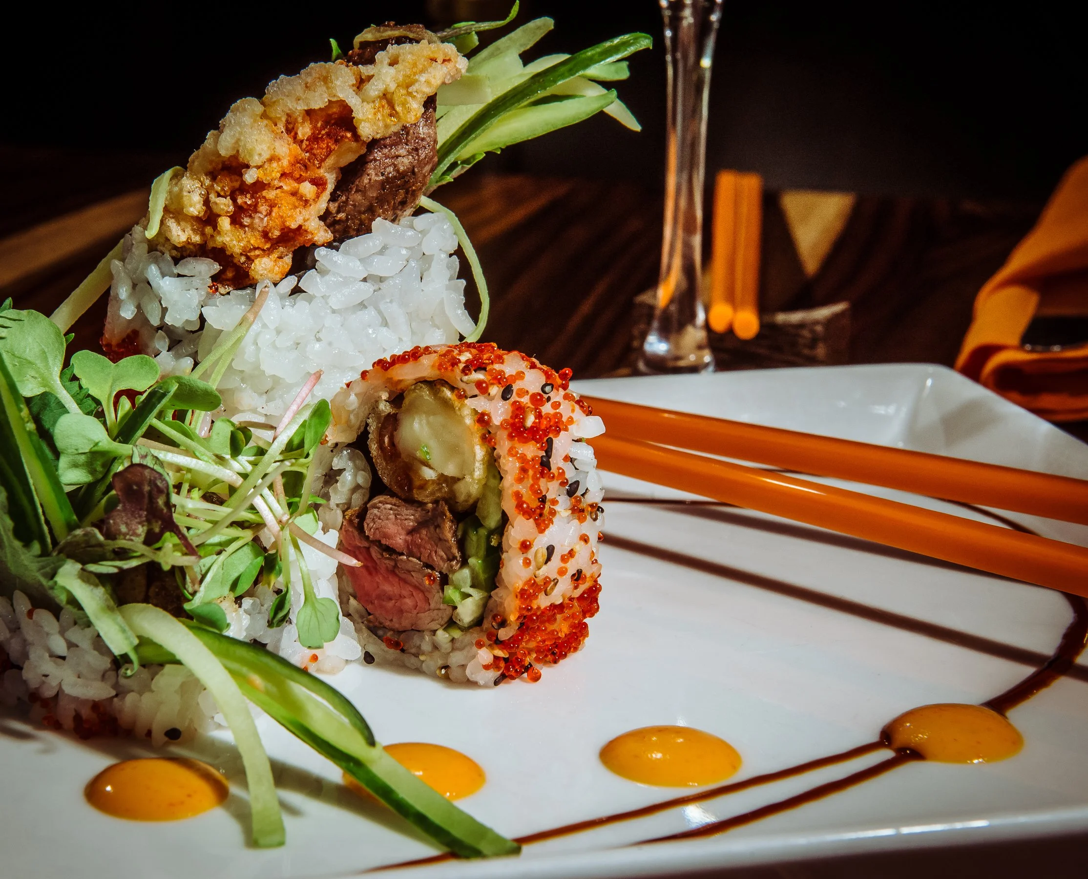 Close-up of a sushi roll with rice, avocado, and beef, garnished with red fish roe, accompanied by various greens, soybean sprouts, and decorative sauces on a white plate.