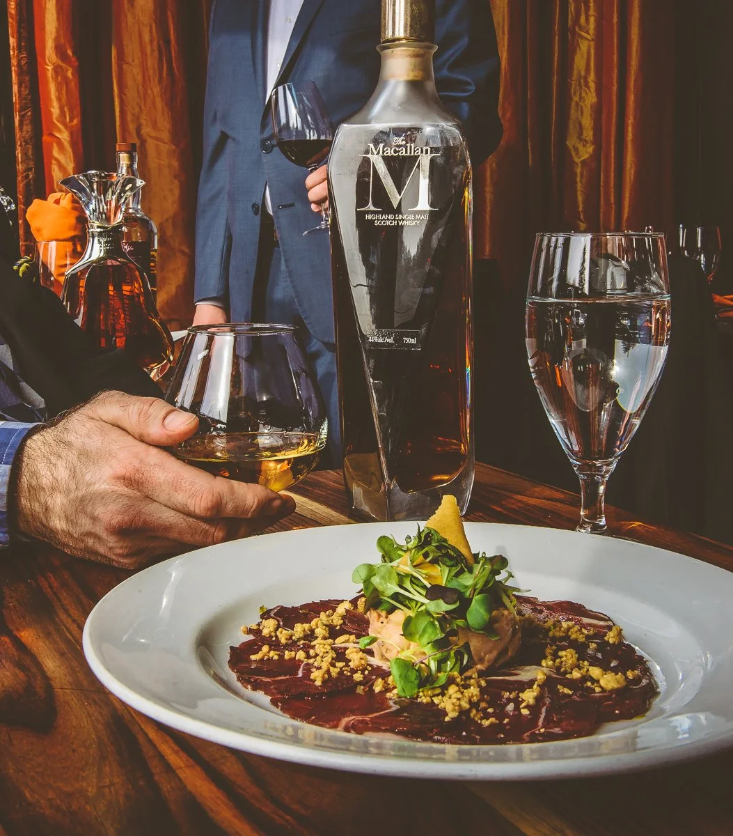 A person in a suit holding a glass of whiskey at a dinner table with a plated dish, a bottle of whisky, a glass of water, and a waiter in the background.