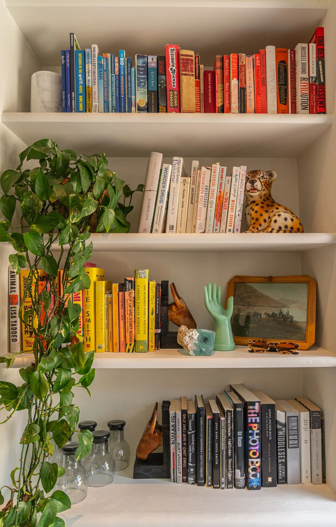 A white bookshelf with four shelves containing books, decorative items, and plants. The top shelf has a marble cylindrical object and books with red, blue, and black covers. The second shelf has a green leafy plant, a sculpture of a leopard, and book