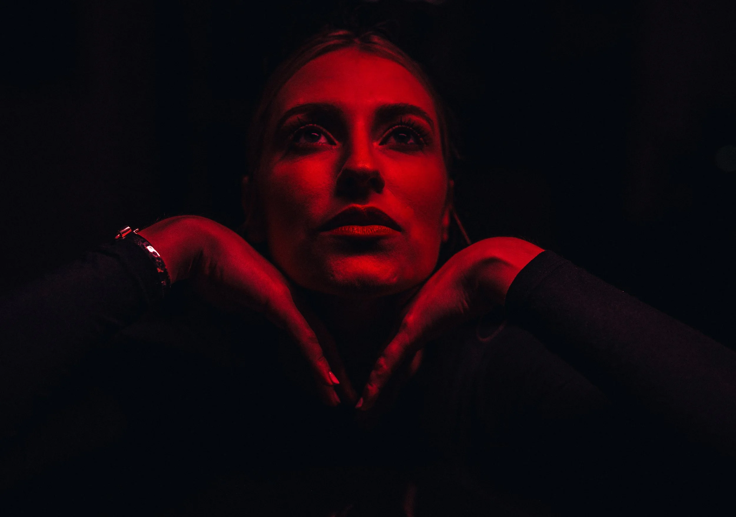 A woman with blonde hair is resting her chin on her hands, which are placed on a dark surface. She is illuminated by red lighting, with a dark background.