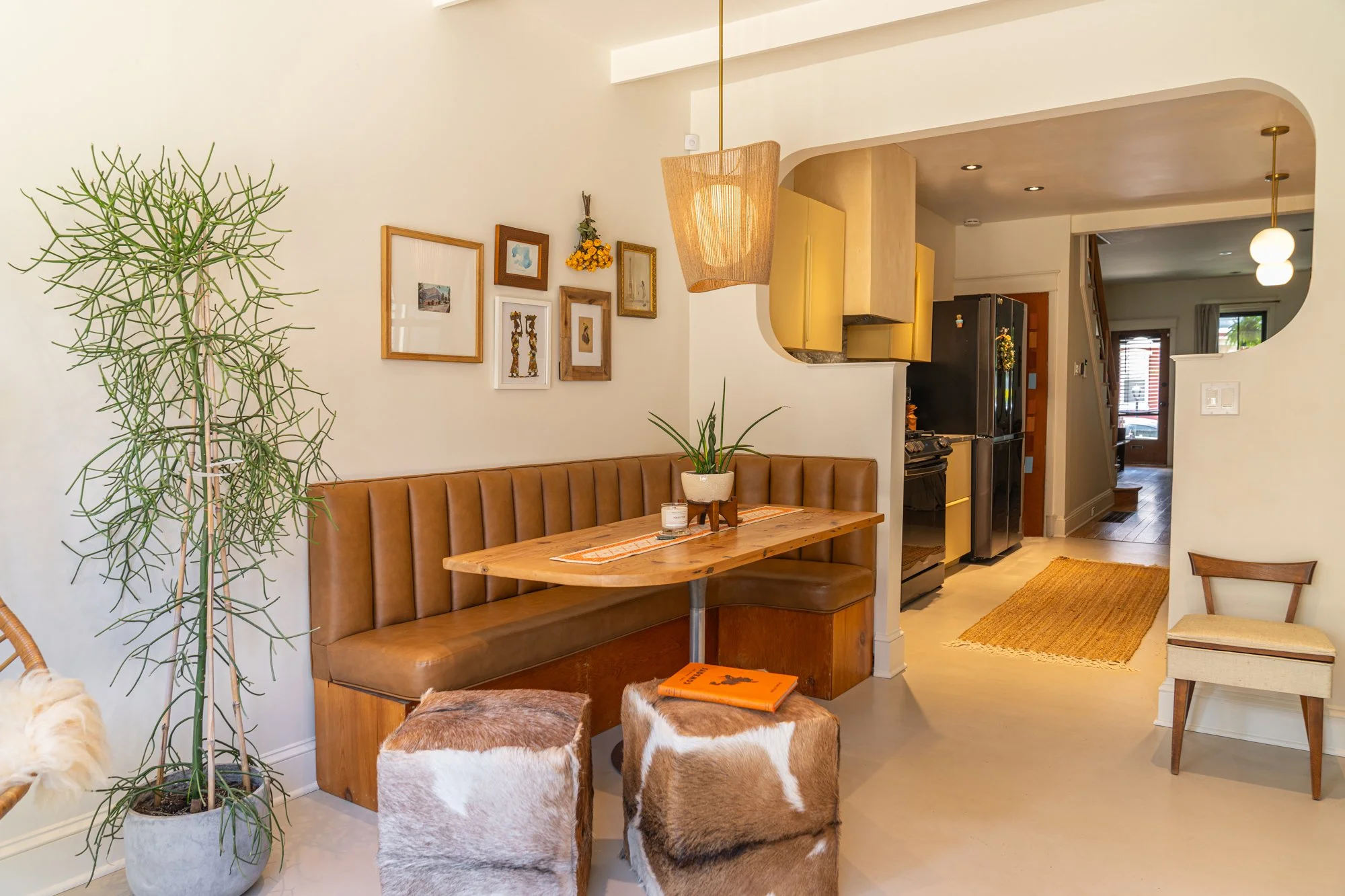 Dining nook with brown leather bench, wooden table, and cowhide ottomans, adjacent to a kitchen with yellow cabinets and black appliances, decorated with framed artwork and potted plants.