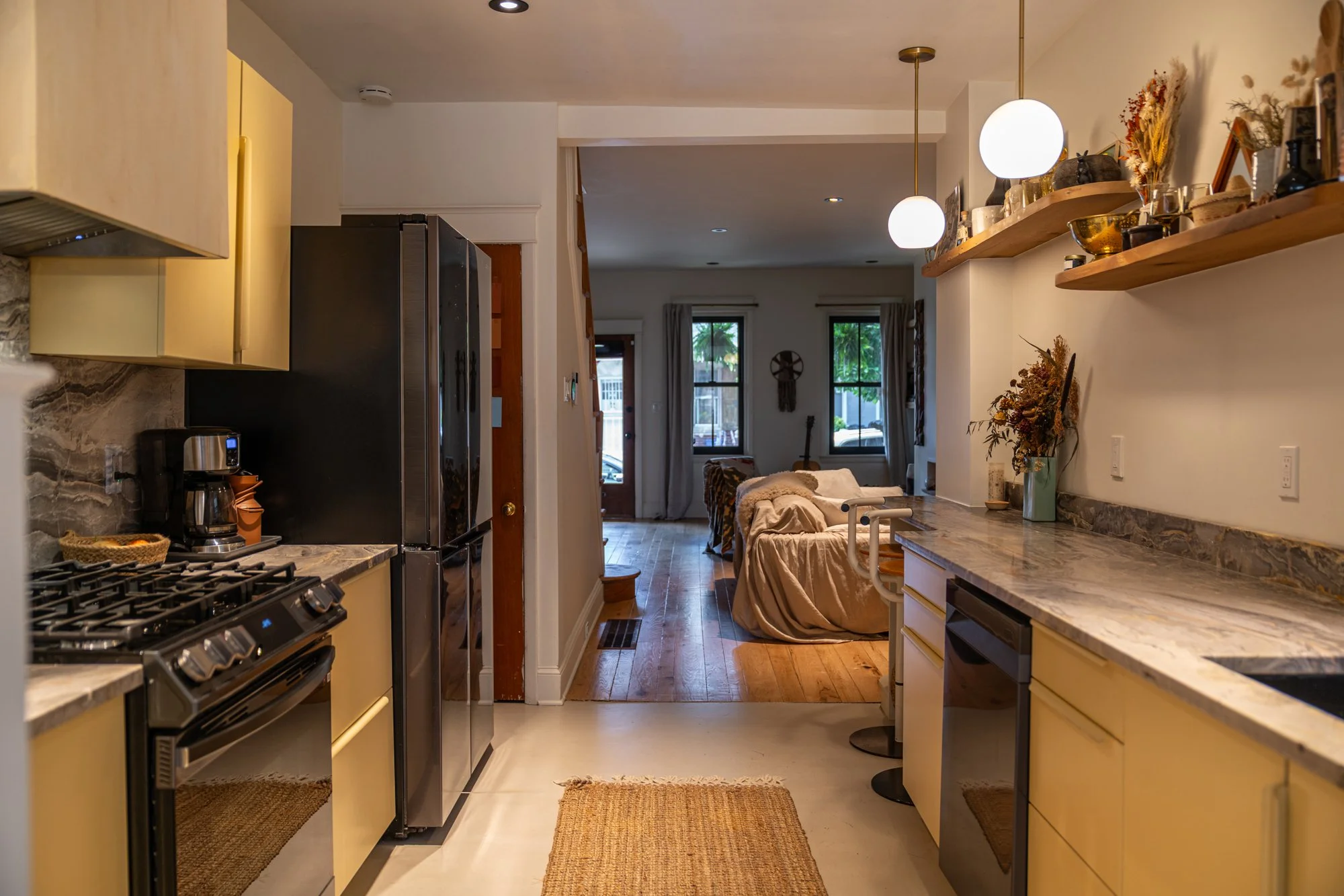 View of a cozy living room seen from a kitchen with yellow cabinets, granite countertops, and modern appliances, featuring a beige couch and windows with curtains, decorated with plants and art.