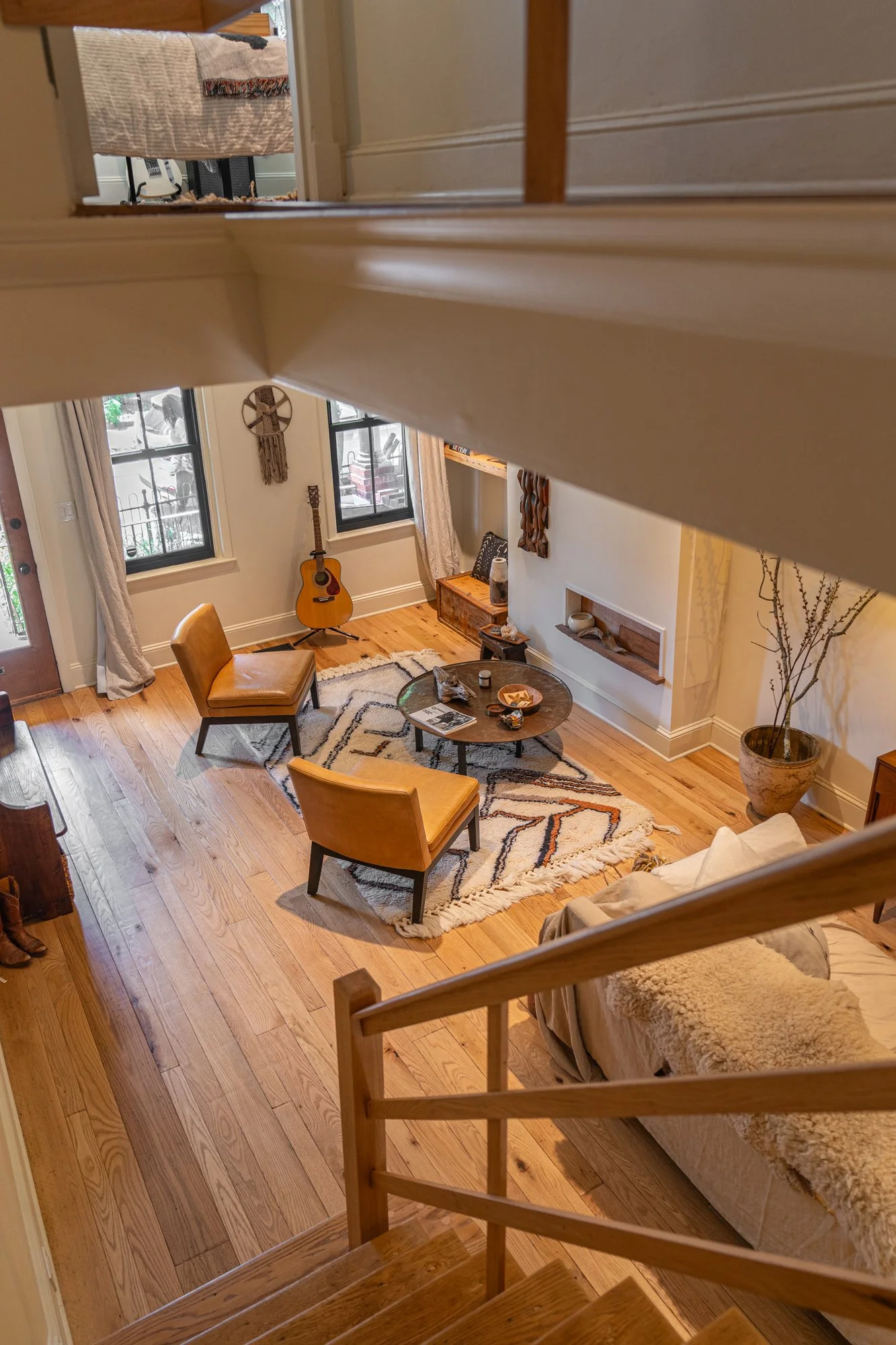 A cozy living room with hardwood floors, a round coffee table, two yellow chairs, a beige sofa with a matching throw blanket, a guitar near the window, and decorative items on the walls and shelves, viewed from the staircase.
