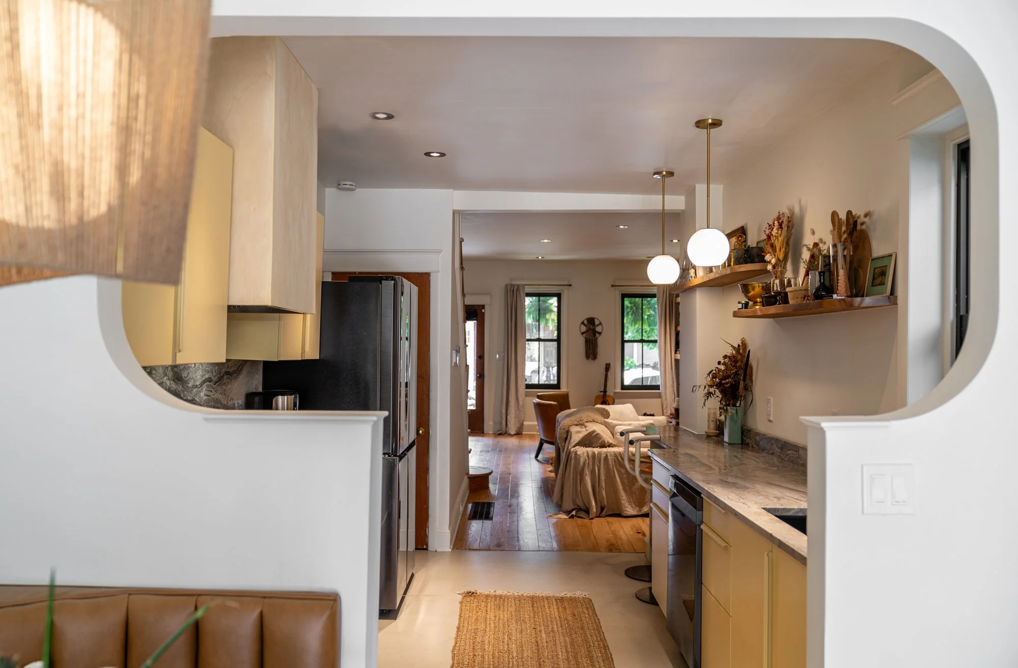 View of a living room and kitchen with an open archway, featuring beige cabinetry, a marble countertop, and decorated shelves with dried flowers and framed pictures, with living room furniture and windows in the background.