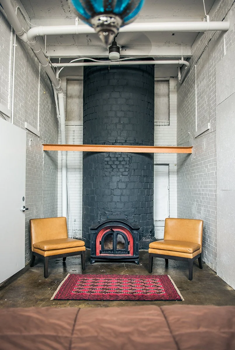 A room with exposed brick walls painted white, a black brick chimney, two tan chairs flanking a small black stove, a red patterned rug, and a wooden shelf mounted on the chimney.