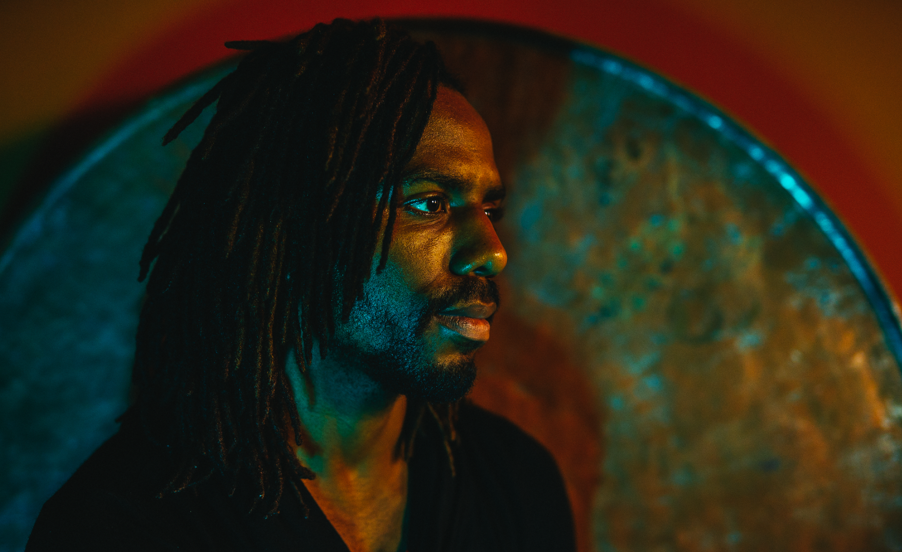 A profile view of a man with shoulder-length dreadlocks, looking to the right, illuminated with multicolored lights, with a large metallic gong in the background.