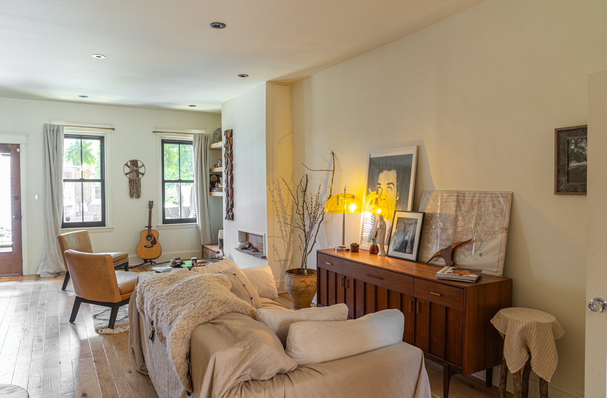 Living room with wooden flooring, a beige sofa with a plush throw, two tan chairs, a wooden sideboard with framed photos and art, a large potted plant, a yellow lamp, and two windows with white curtains.