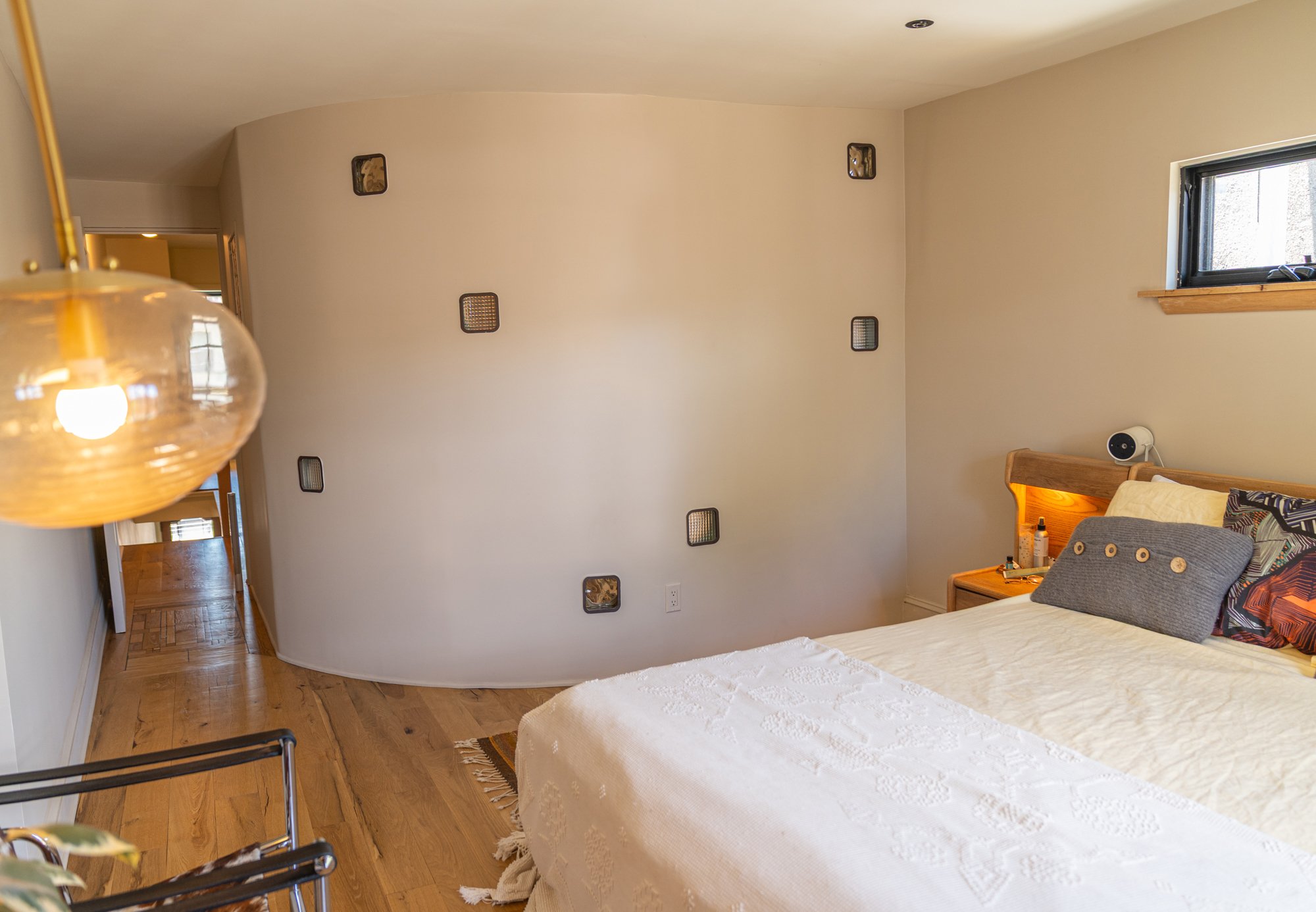 A bedroom with a bed covered in a white lace bedspread, and decorative pillows, a wooden headboard with lighting, a small window on the right, a side table with toiletries, and a textured wall with small square vents.