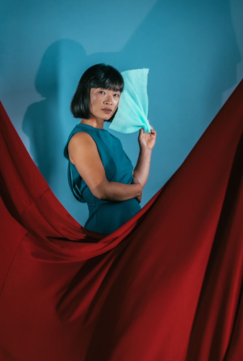 Woman with short black hair wearing a blue dress holding a blue cloth near her face, standing behind red drapes against a blue background.