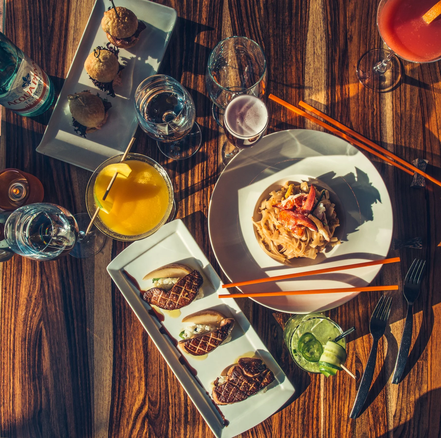A top-down view of a wooden table with various dishes and drinks, including pasta, sliced meats, cocktails, and bottled water.