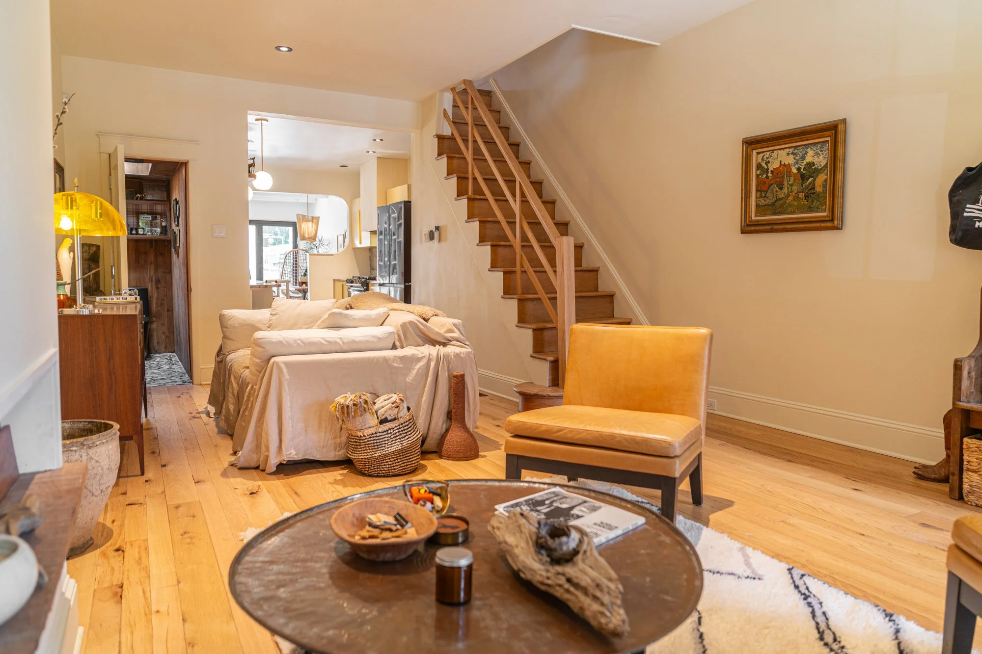 Living room with beige walls, wooden staircase, yellow armchair, couches covered with beige sheets, coffee table with decorative items, and a bright open kitchen in the background.