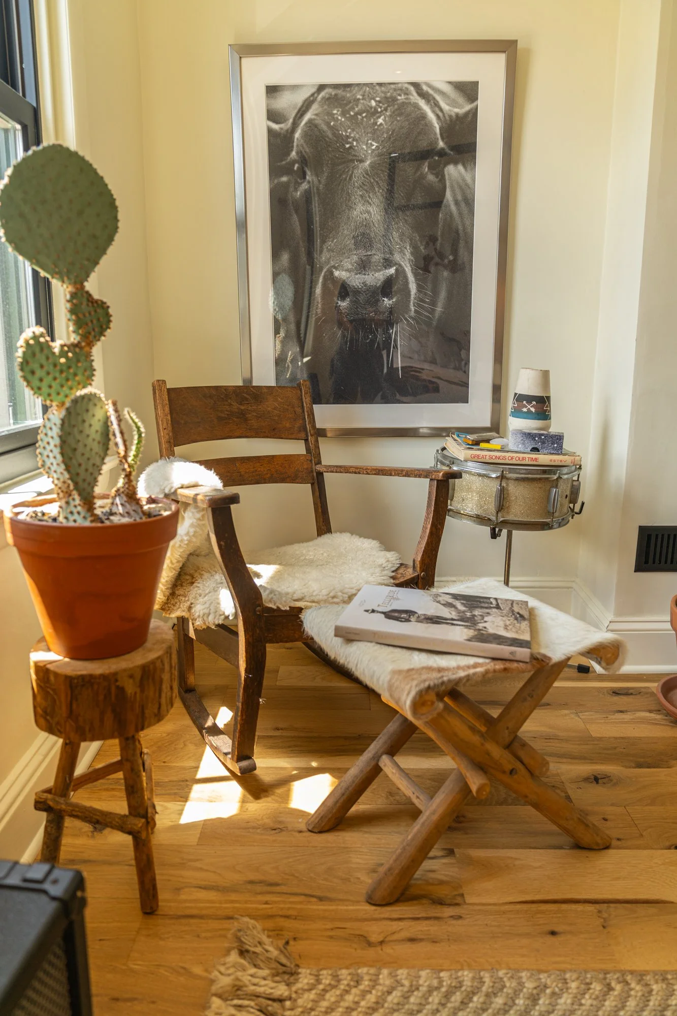 A cozy corner with a wooden armchair, a matching footstool, and a side table decorated with a cactus plant, books, and a lamp. A large black-and-white photograph of a cow hangs on the wall.