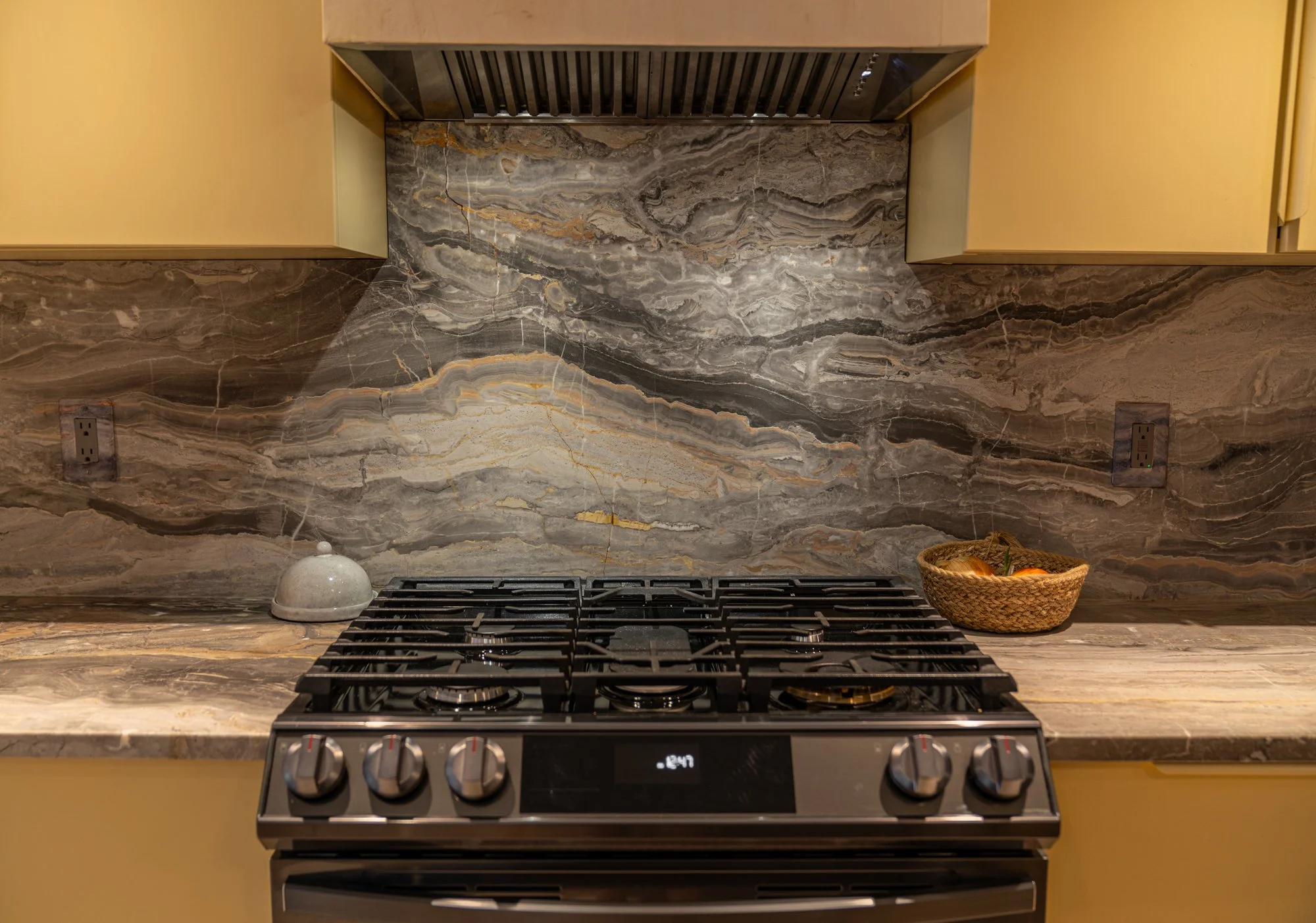 Kitchen stove with a marble backsplash and countertop, with a small ceramic bell on the left and a basket of onions on the right.