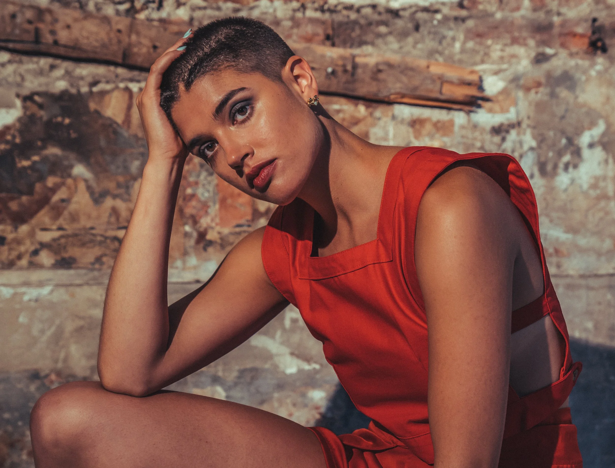 A woman with short braided hair and wearing a red sleeveless top and shorts, sitting against a rustic brick wall.