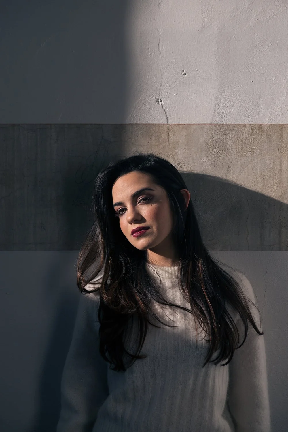 A woman with long dark hair and makeup, wearing a cream-colored sweater, stands against a textured wall with sunlight casting shadows on her face.