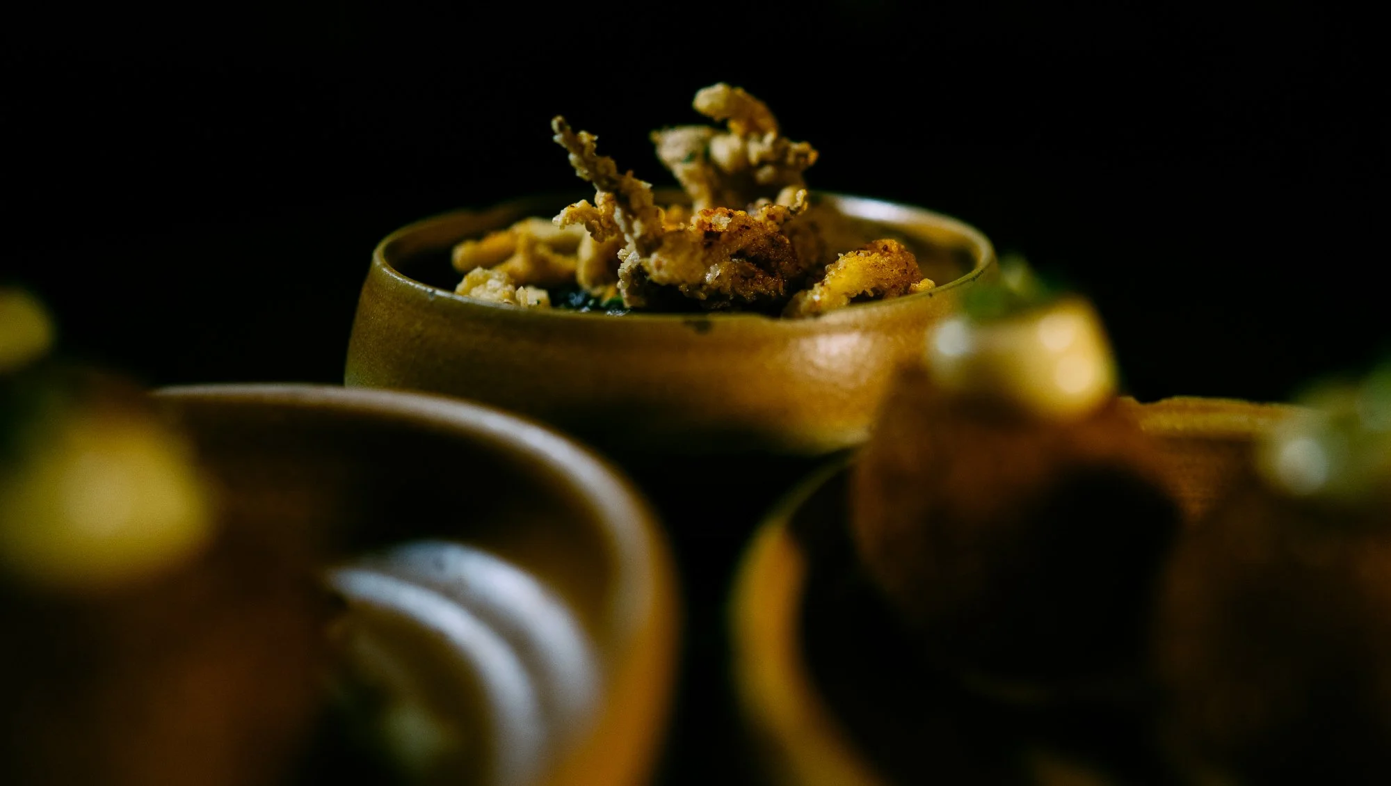 Close-up of a small wooden bowl filled with fried insects against a dark background, with other bowls partially visible around it.