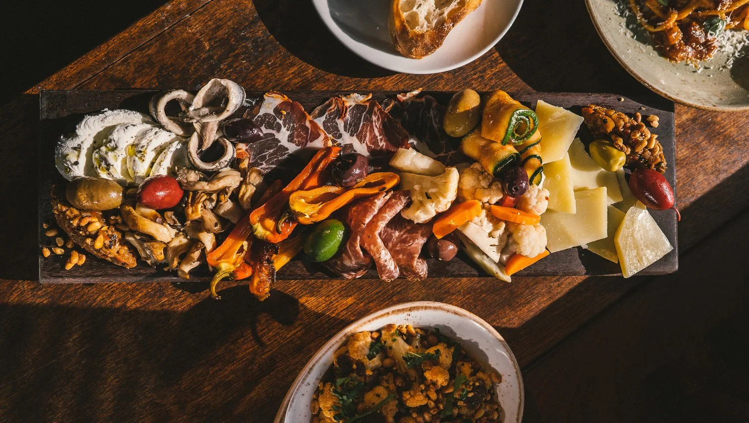 Charcuterie board with assorted cheeses, cured meats, roasted vegetables, and olives, accompanied by a bowl of seasoned chickpeas or lentils and some bread slices on a dark wooden table.