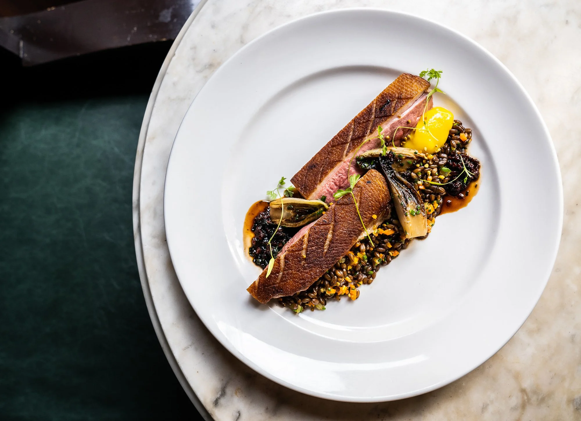 A dish with a seared piece of meat, topped with microgreens, served on a bed of lentils, with artichoke hearts and a yellow sauce, presented on a white plate.