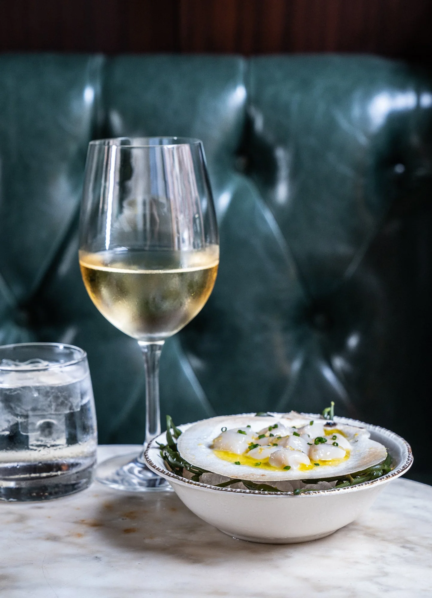 A glass of white wine, a glass of water, and a bowl of eggs with greens on a marble table against a dark green upholstered background.
