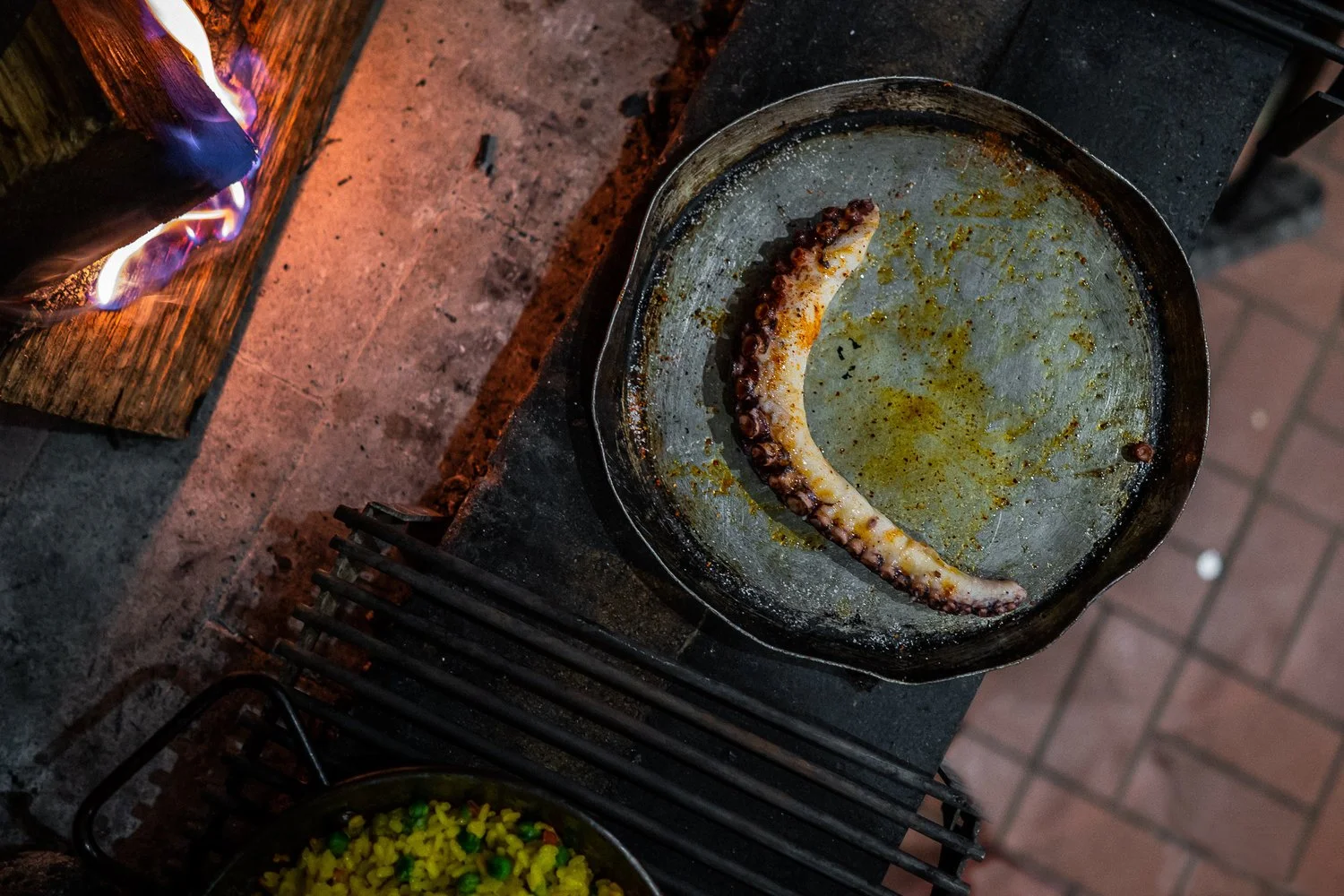 Octopus tentacle being grilled on a round metal grill with olive oil and spices, beside a wood fire stove with blue flames.