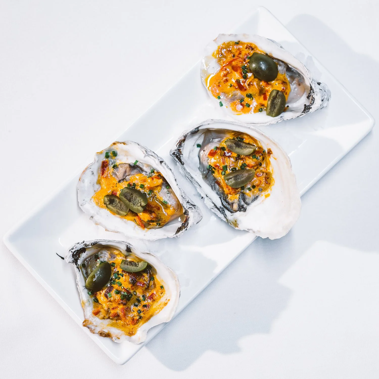 Five baked oysters topped with cheese, bacon bits, chopped chives, and a jalapeño slice on each, arranged on a white rectangular plate.