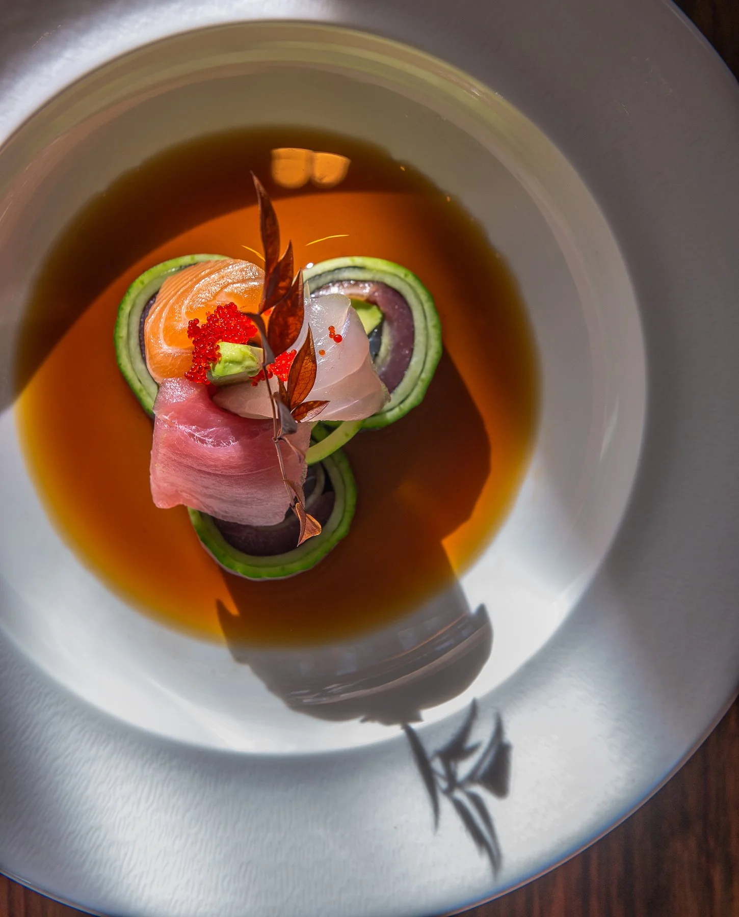 A gourmet dish featuring thinly sliced fresh fish, salmon, and vegetables like cucumber, all artfully arranged in a dark broth and garnished with red fish roe and delicate edible leaves.