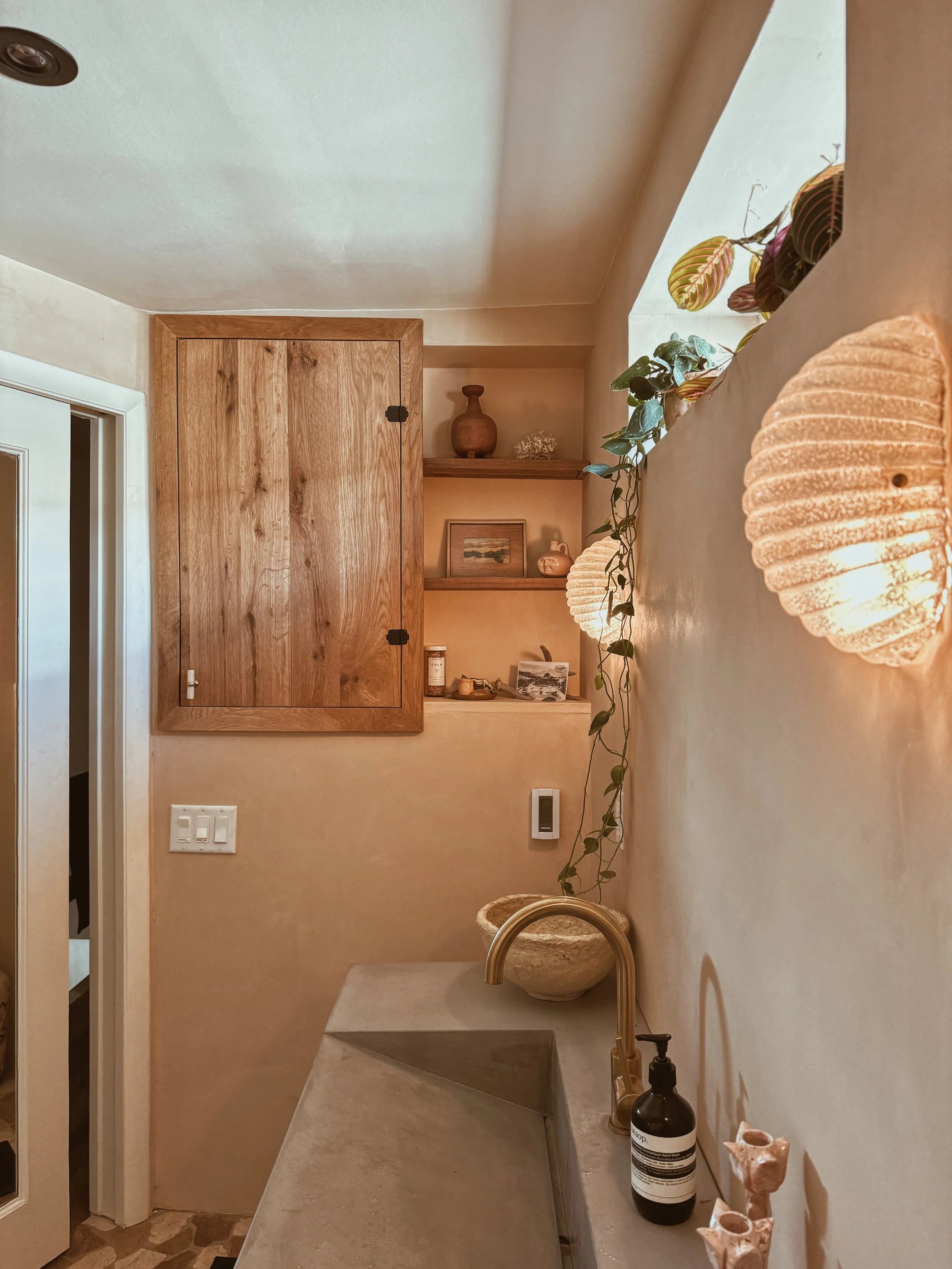 Interior of a cozy room with beige walls, a concrete countertop sink with a gold faucet, a black soap dispenser, and decorative items. There are wooden shelves with vases and artwork, a wooden cabinet door, a hanging plant, and textured wall lights.