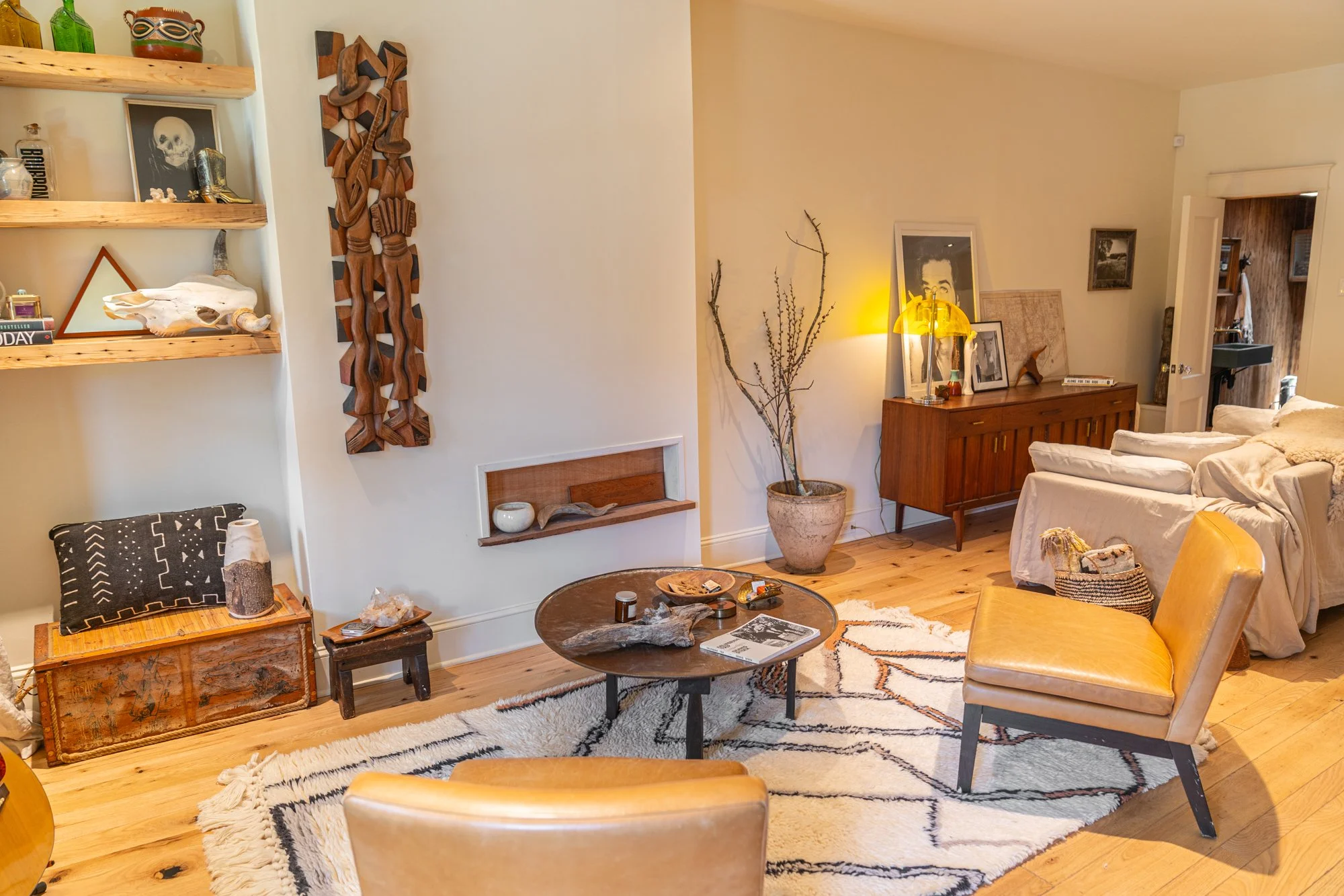 A cozy living room with wooden flooring, a white textured area rug, two tan armchairs, a round coffee table with decorative items, a wooden sideboard with framed photos and a yellow lamp, a large potted branch, and wall art. Shelves with decorative o