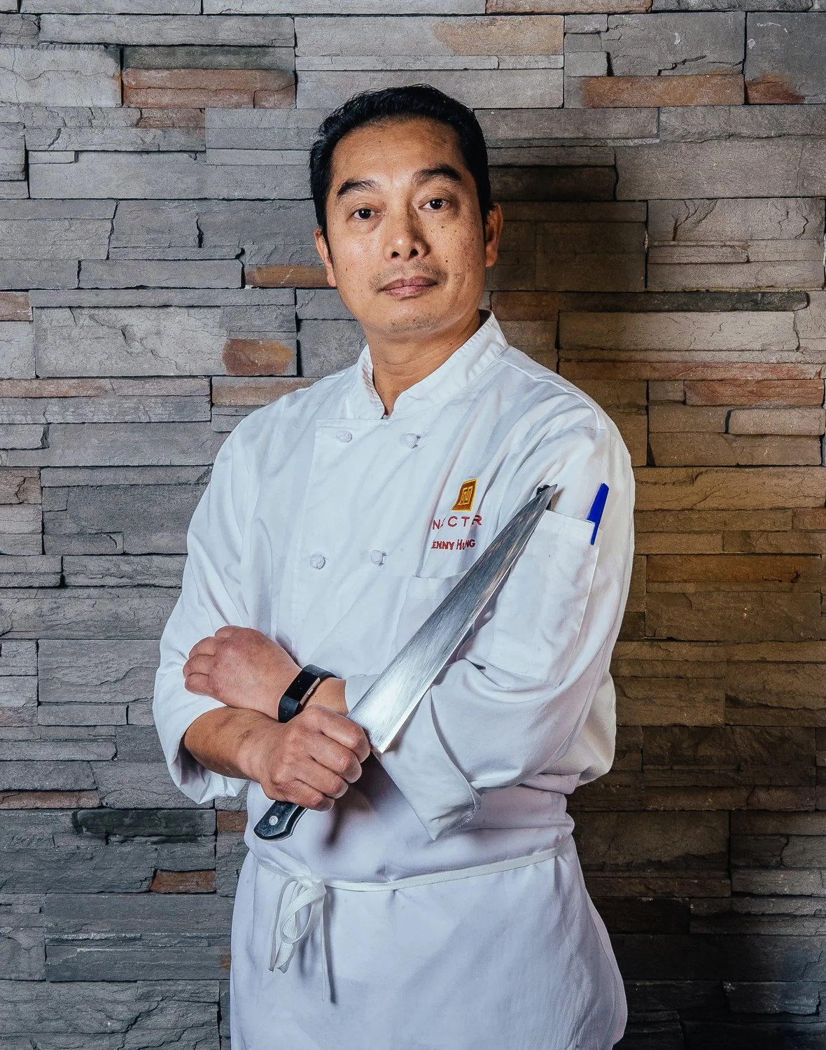 A male chef dressed in a white chef coat, holding a large kitchen knife across his chest, standing in front of a textured gray stone wall.
