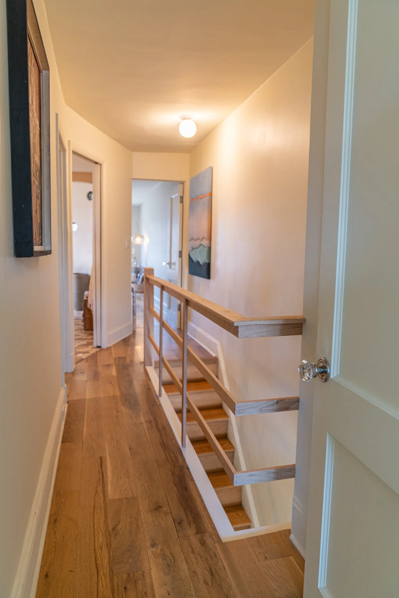 Hallway with wooden flooring, artwork on the walls, and a staircase with a wooden handrail leading down.