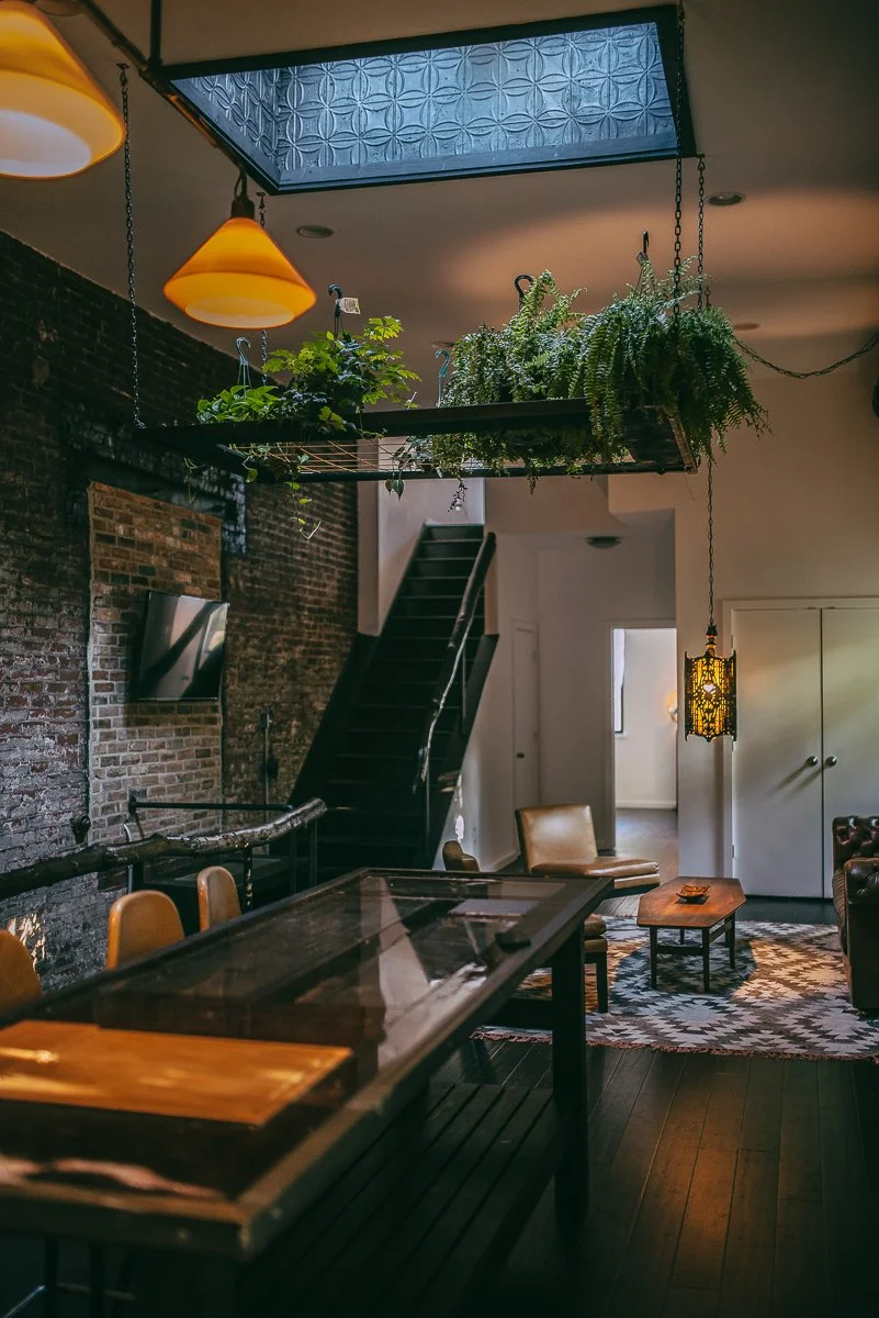 Interior of a cozy living space featuring a brick wall, a hanging plant shelf with lush greenery, a black staircase, a flat-screen TV, a wooden coffee table, leather chairs, pendant lighting, and a patterned rug.