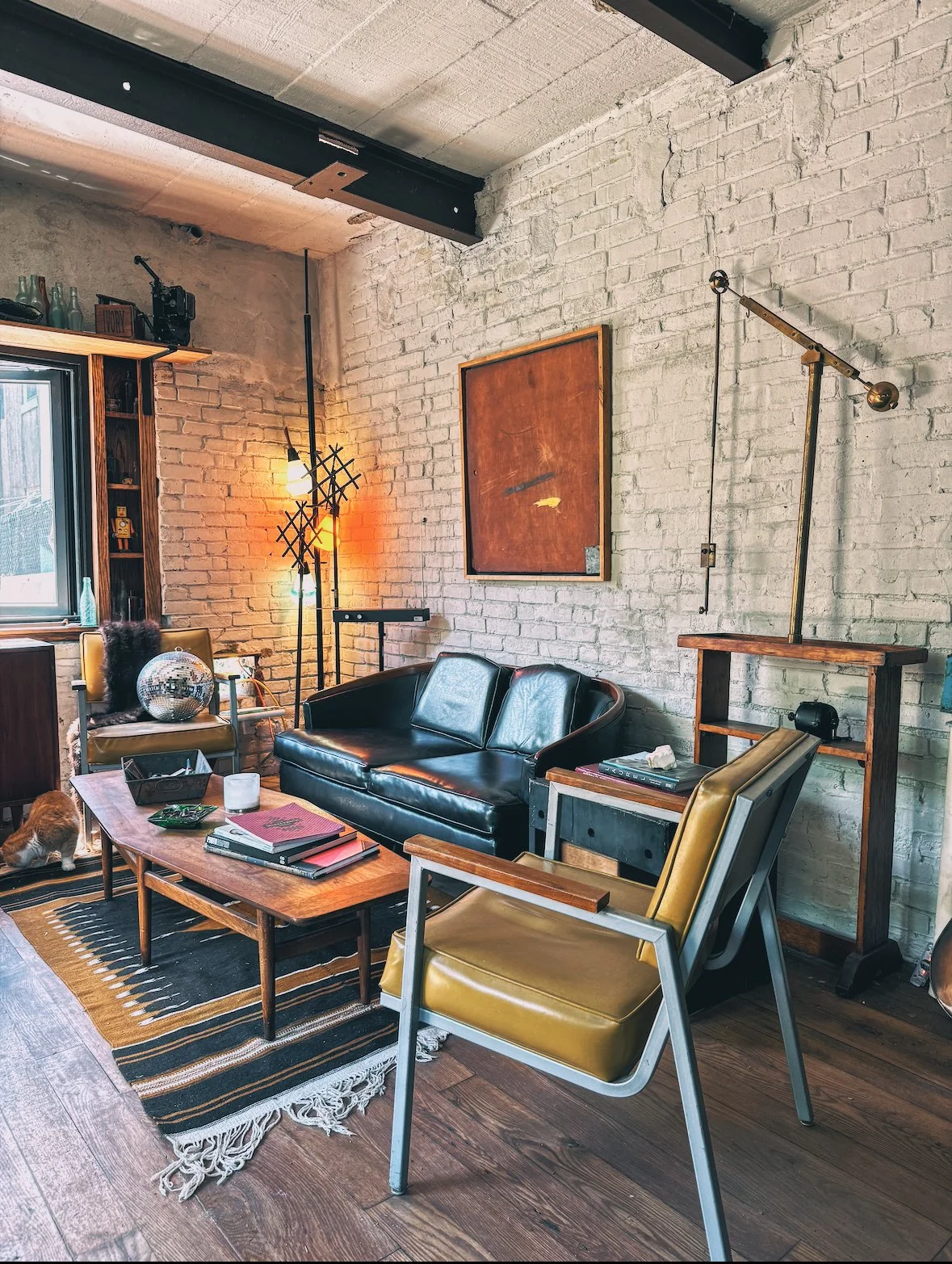 Living room with white brick walls, a black leather sofa, yellow and gray chairs, a wooden coffee table, and an art piece on the wall. A window with natural light, a black metal floor lamp, and decorative items like a disco ball and plants are visibl