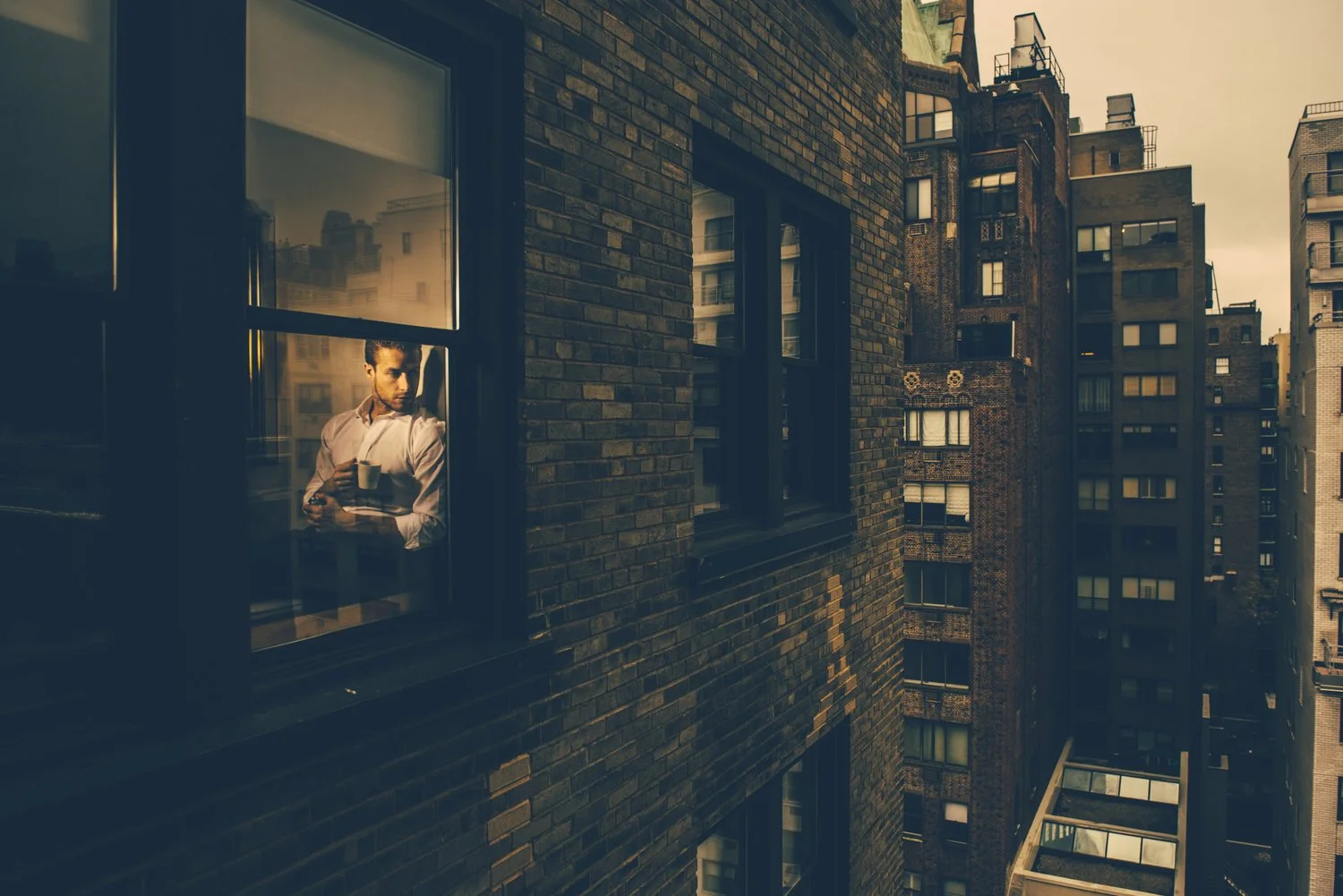 A man holding a mug is looking out of a window in an apartment building during sunset. The building has a brick exterior, and surrounding high-rise buildings are visible in the background.