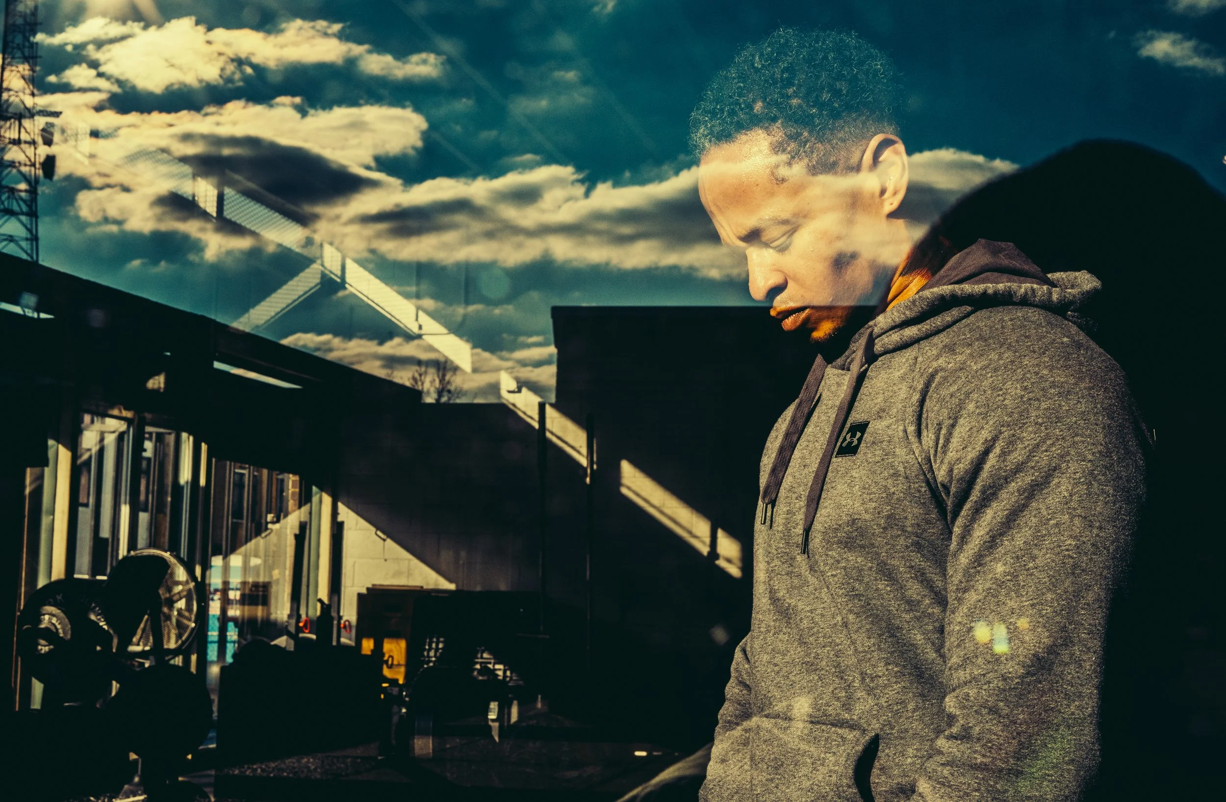 A man in a gray hoodie standing outside, viewed through a glass window that reflects the sky with clouds and a sunset or sunrise, along with structures like a building and a bridge or crane.