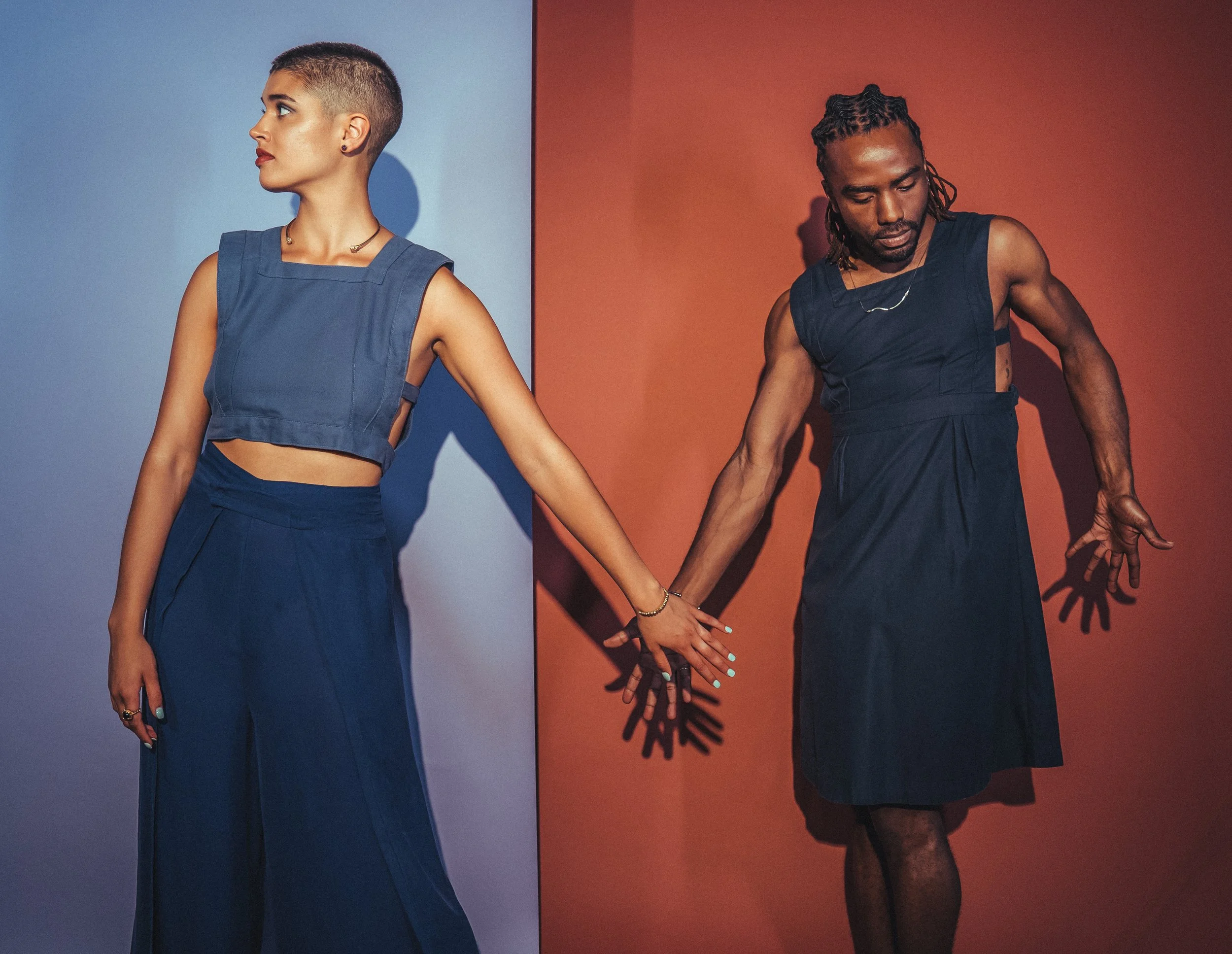 Two people, one woman with short hair and makeup, and one man with dreadlocks, are holding hands against contrasting backgrounds, blue on the left and red on the right.