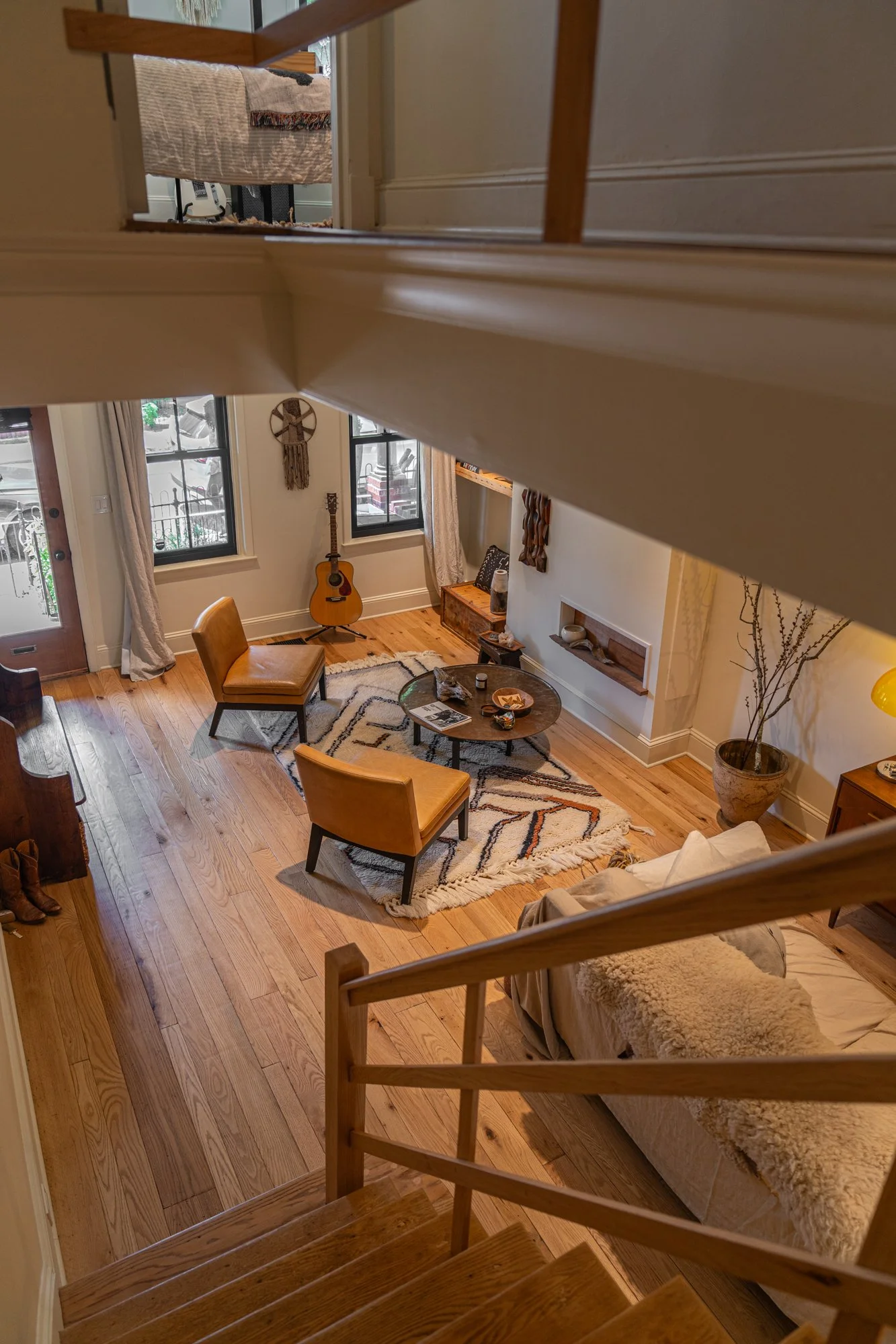 Living room viewed from the staircase, featuring a sofa, two chairs, a round coffee table, a guitar, a rug, large windows with curtains, and various decorative items.