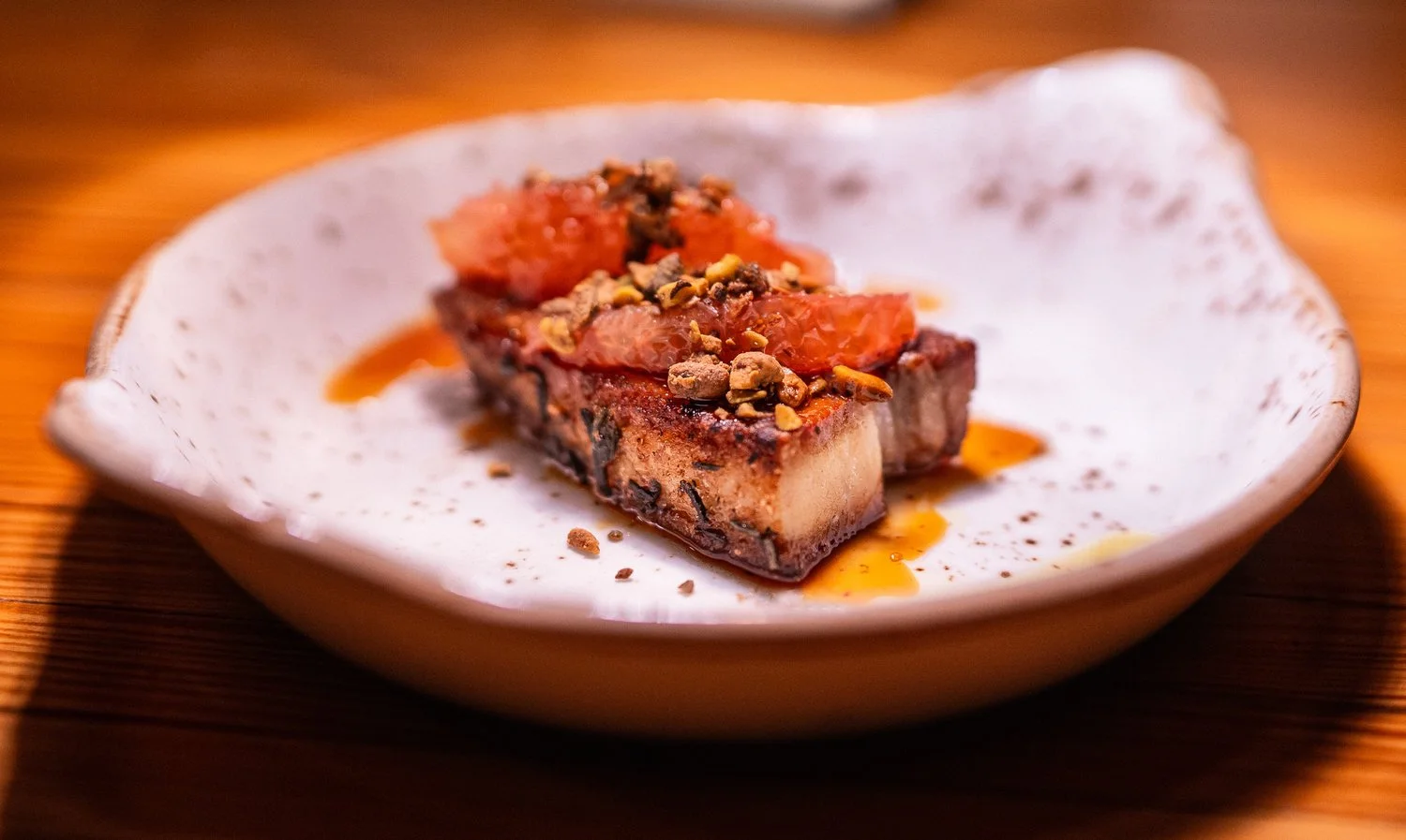 A dessert with a chocolate base topped with tomato slices and crushed nuts, served on a white ceramic plate with a wooden surface background.