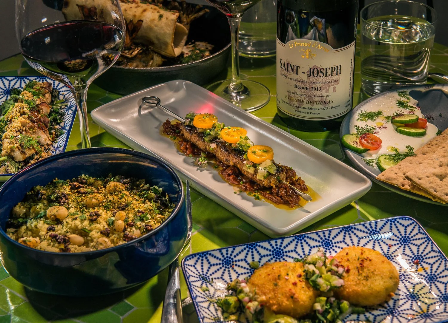 A table set with various dishes including a bowl of couscous with chickpeas and herbs, a plate of falafel with chopped onions and herbs, a skewered grilled meat dish topped with vegetables, a plate of cucumber and tomato salad with herbs, and a bottl