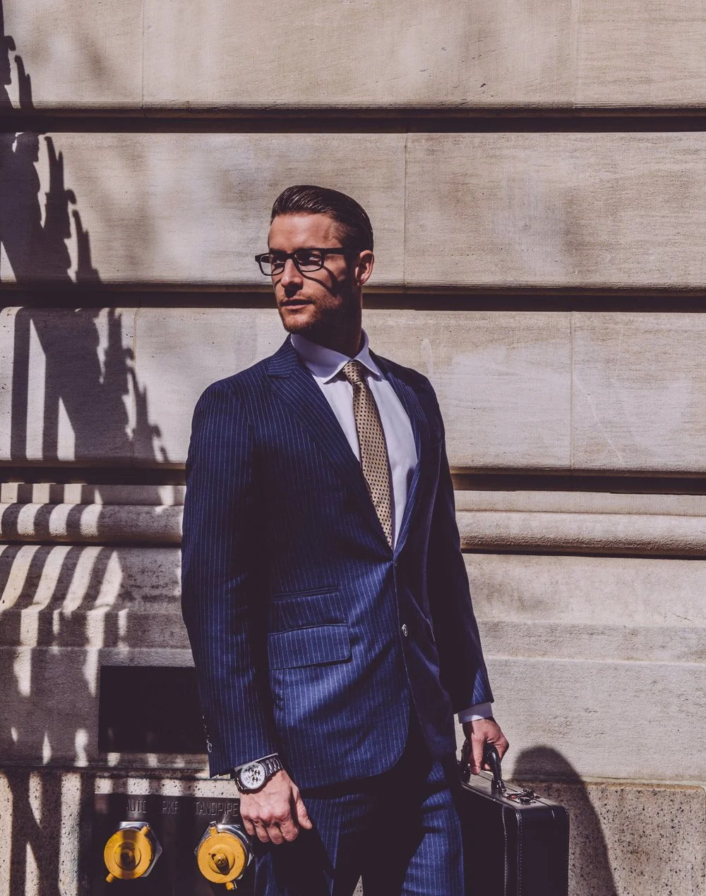 A man in a pinstripe suit and glasses standing against a stone wall with shadows cast on it, holding a briefcase.