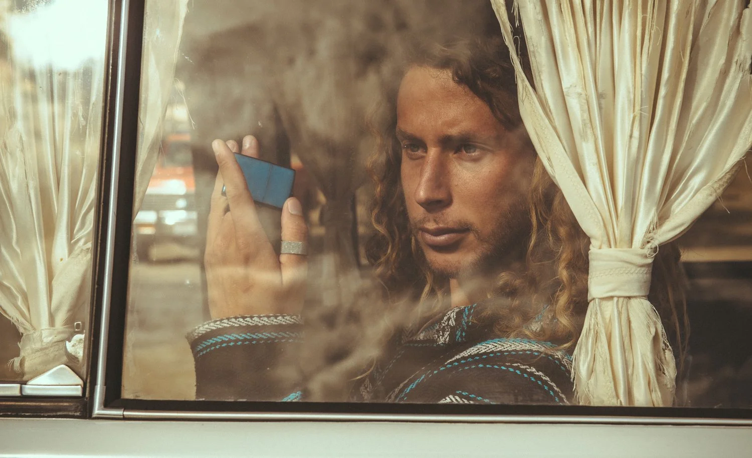 A man with long curly hair and tan complexion looking out of a vehicle window, holding a mobile phone. The window has cream-colored curtains tied back.