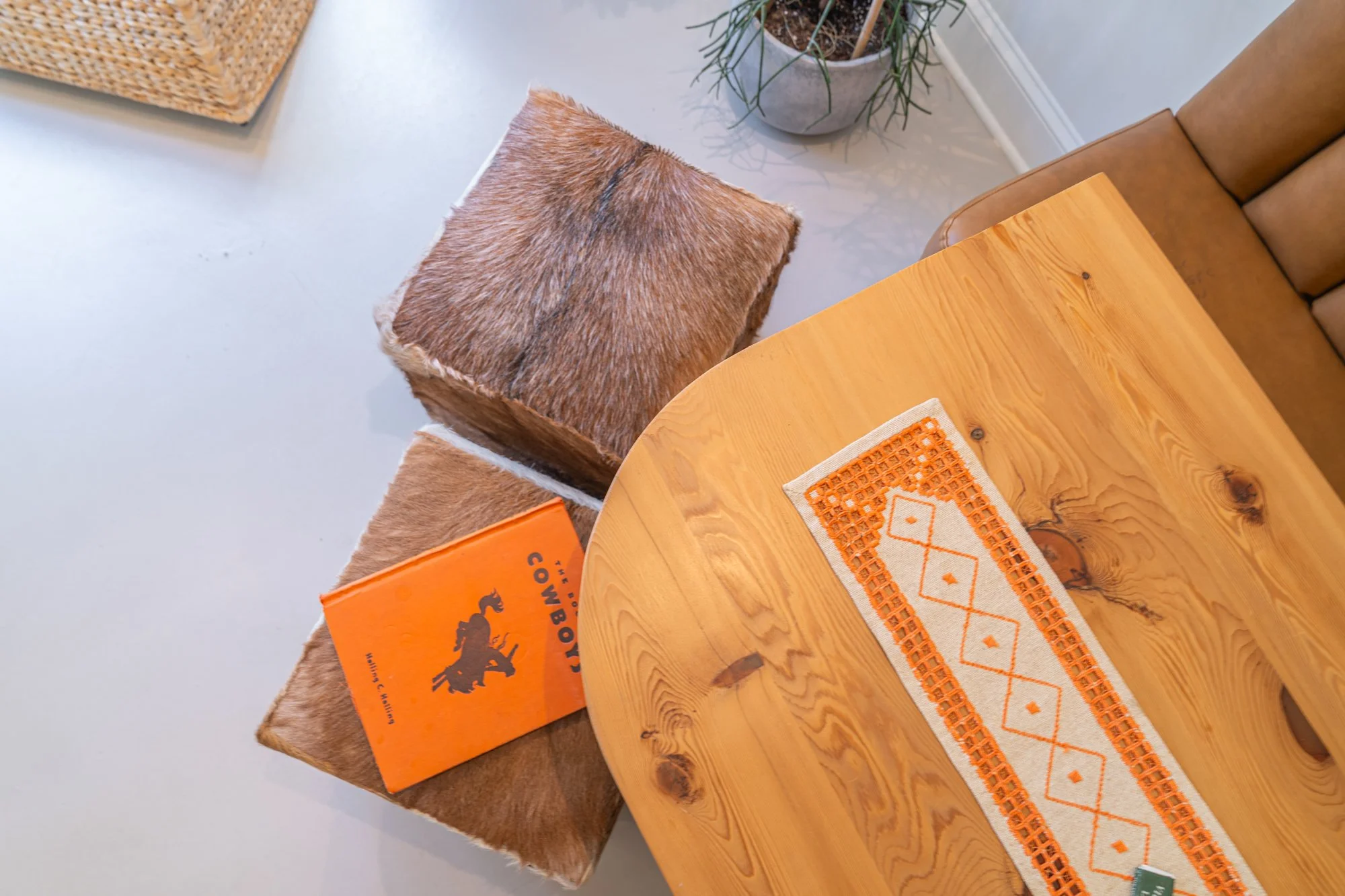 A wooden dining table with orange and white patterned table runner, surrounded by a leather chair and fuzzy cube-shaped stools, with a plant and a box with an orange and black label in the background.