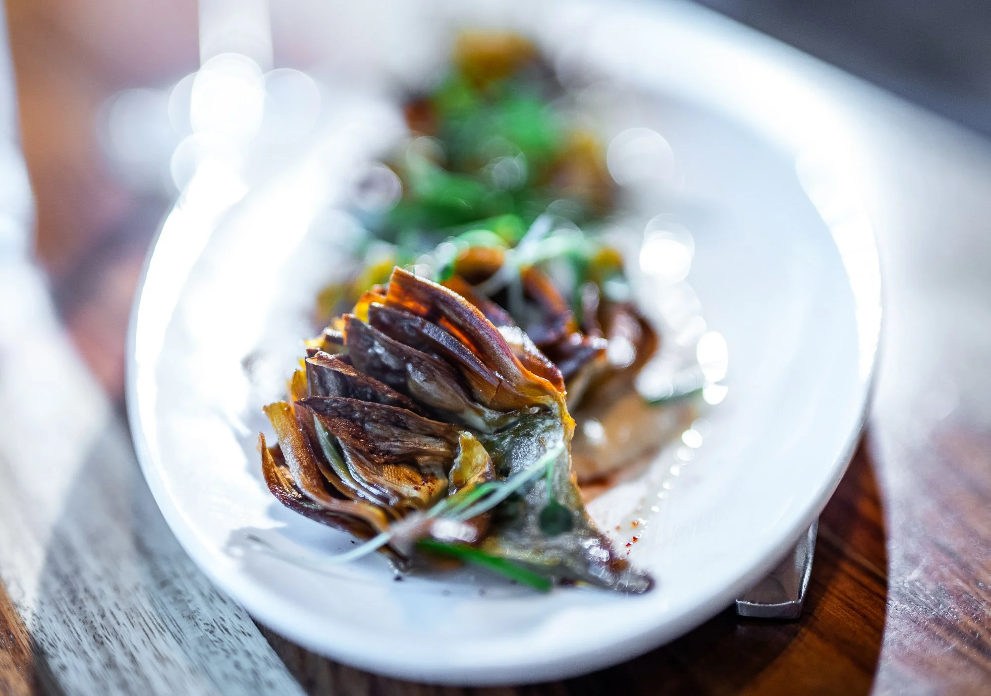 Close-up of a plate with grilled or roasted artichoke heart garnished with herbs, served on a white dish on a wooden table.
