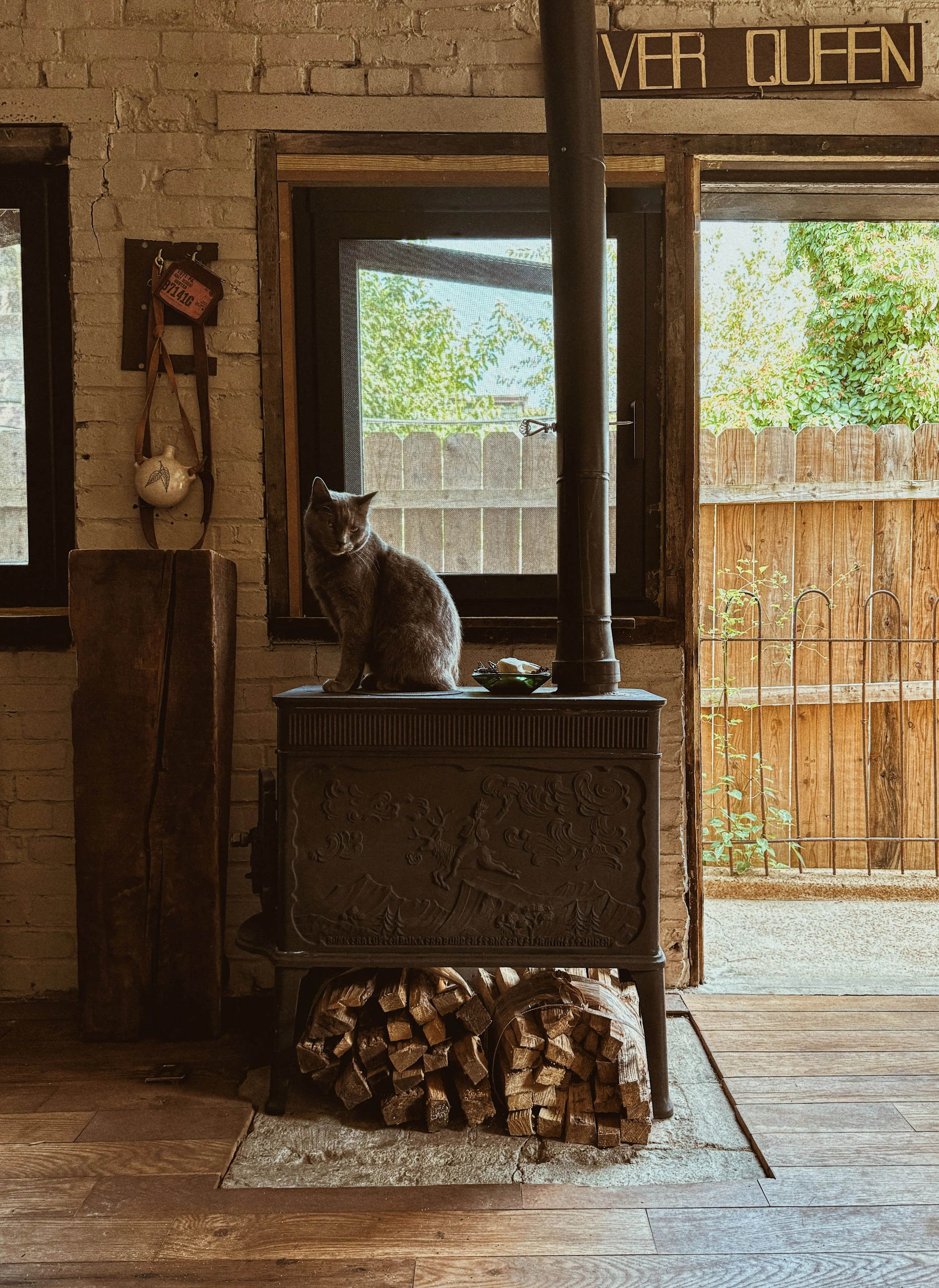 A cat sitting on a vintage cast iron stove in a rustic kitchen with exposed brick walls, wood flooring, and a window looking outside to a wooden fence and greenery. There are logs of firewood beneath the stove, a sign on top of the window frame that 