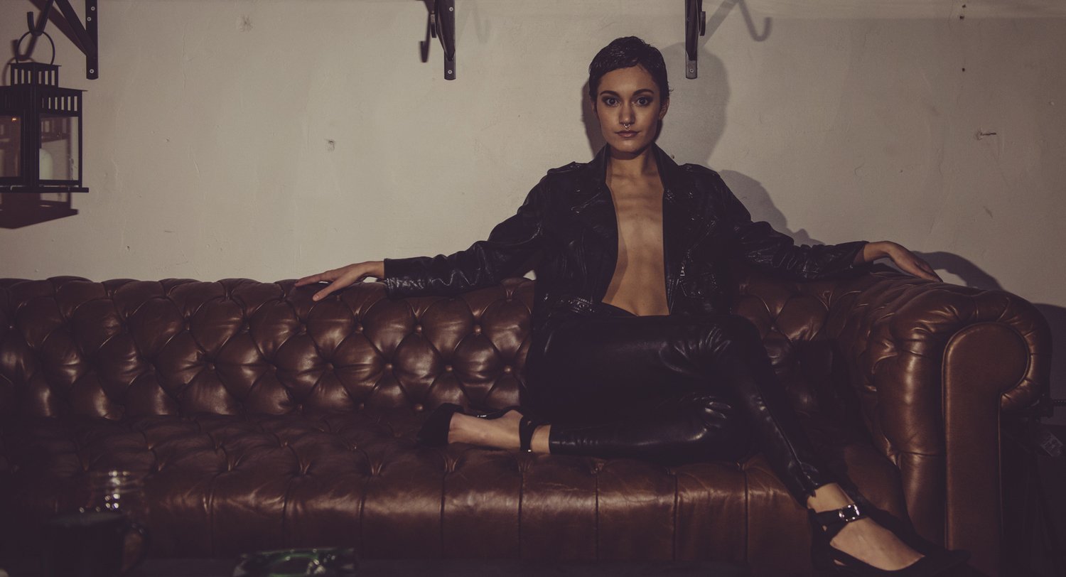 A woman with short dark hair and a septum piercing, wearing black leather pants and a black leather jacket, sits on a brown leather couch in a dimly lit room with white walls and wall-mounted hooks.