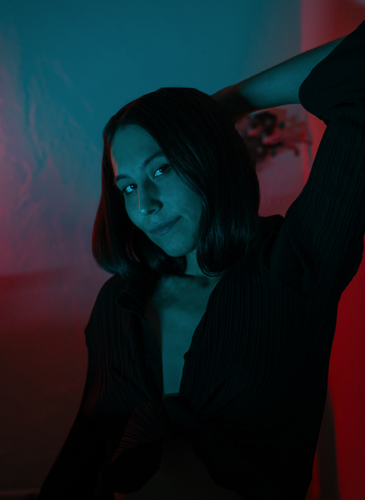 A woman with shoulder-length dark hair posing with one arm raised behind her head, wearing a dark striped shirt, illuminated with colorful red and blue lighting in a dimly lit setting.
